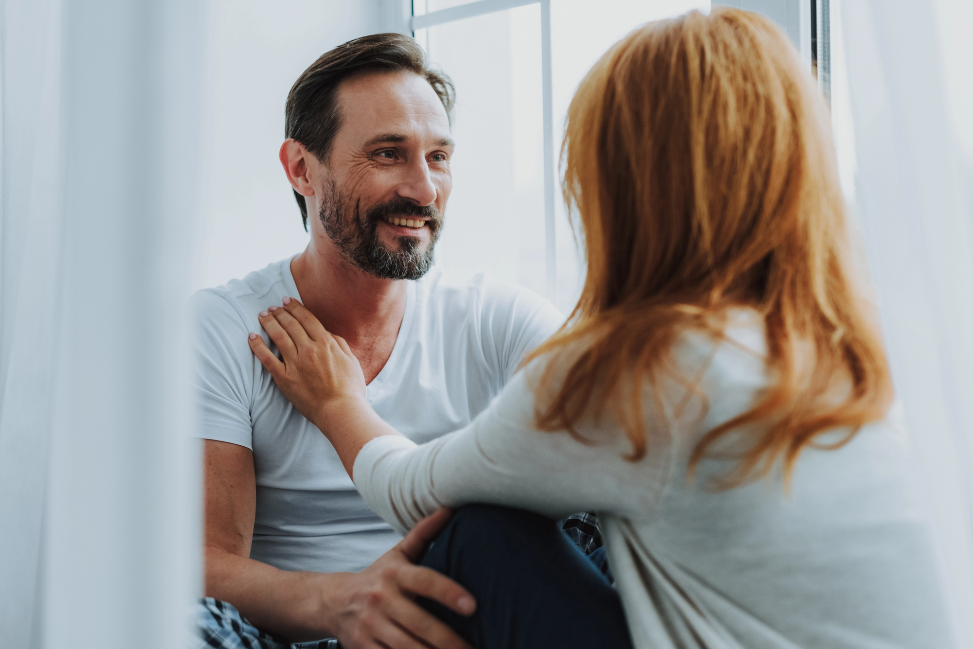 A man with a beard and a woman with red hair sit by a window, smiling and engaged in conversation. The man wears a white t-shirt and the woman has her hand on his shoulder. Natural light filters through sheer curtains.