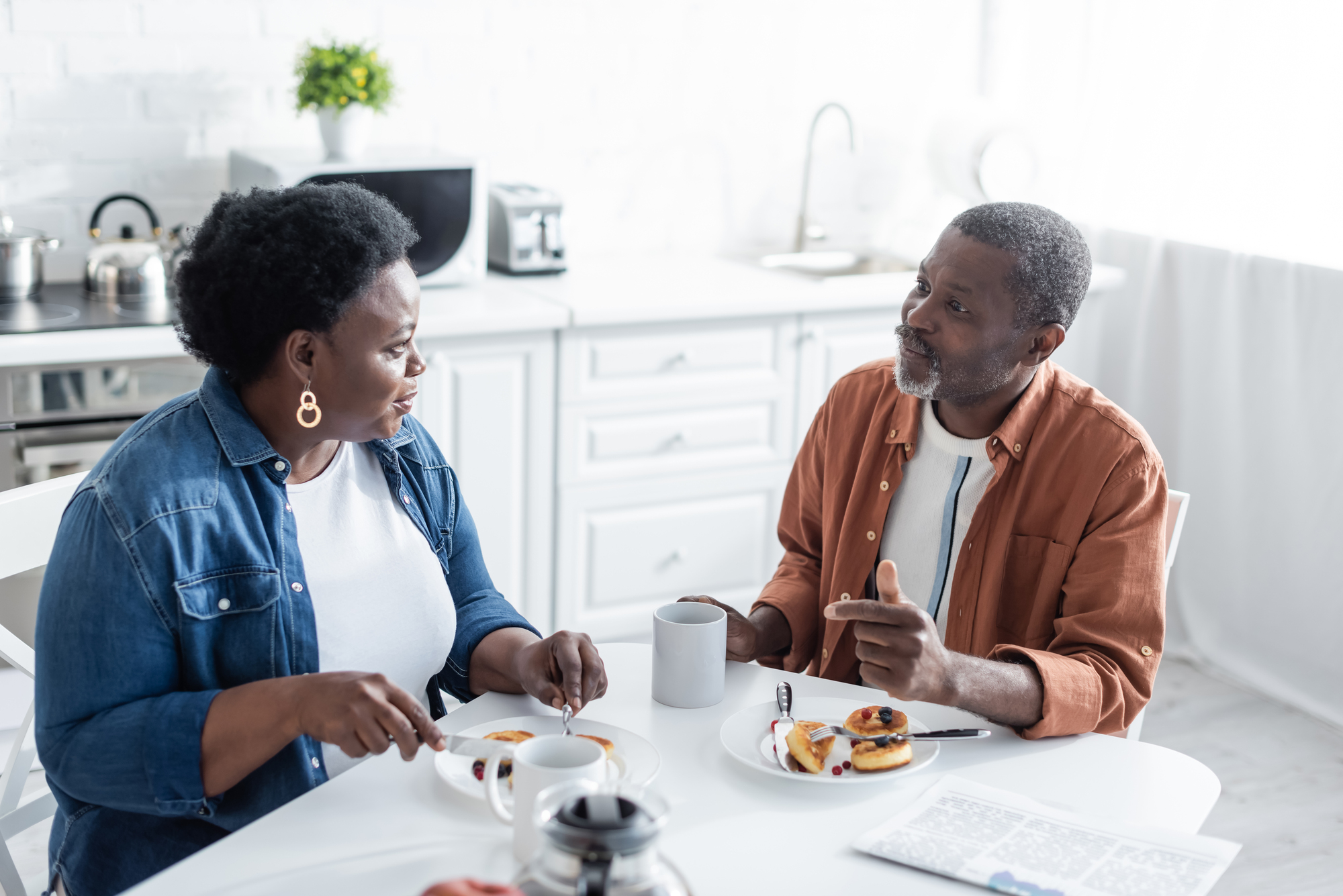 A middle-aged couple sits at a kitchen table, enjoying breakfast. The woman wears a blue jacket, and the man a brown one. They have coffee and pastries. The room is bright and modern, with white cabinets and a bit of greenery in the background.