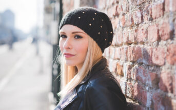 A woman with long blonde hair wearing a black beanie and leather jacket leans against a brick wall. She gazes sideways with a calm expression. The background is blurred, suggesting an urban setting.