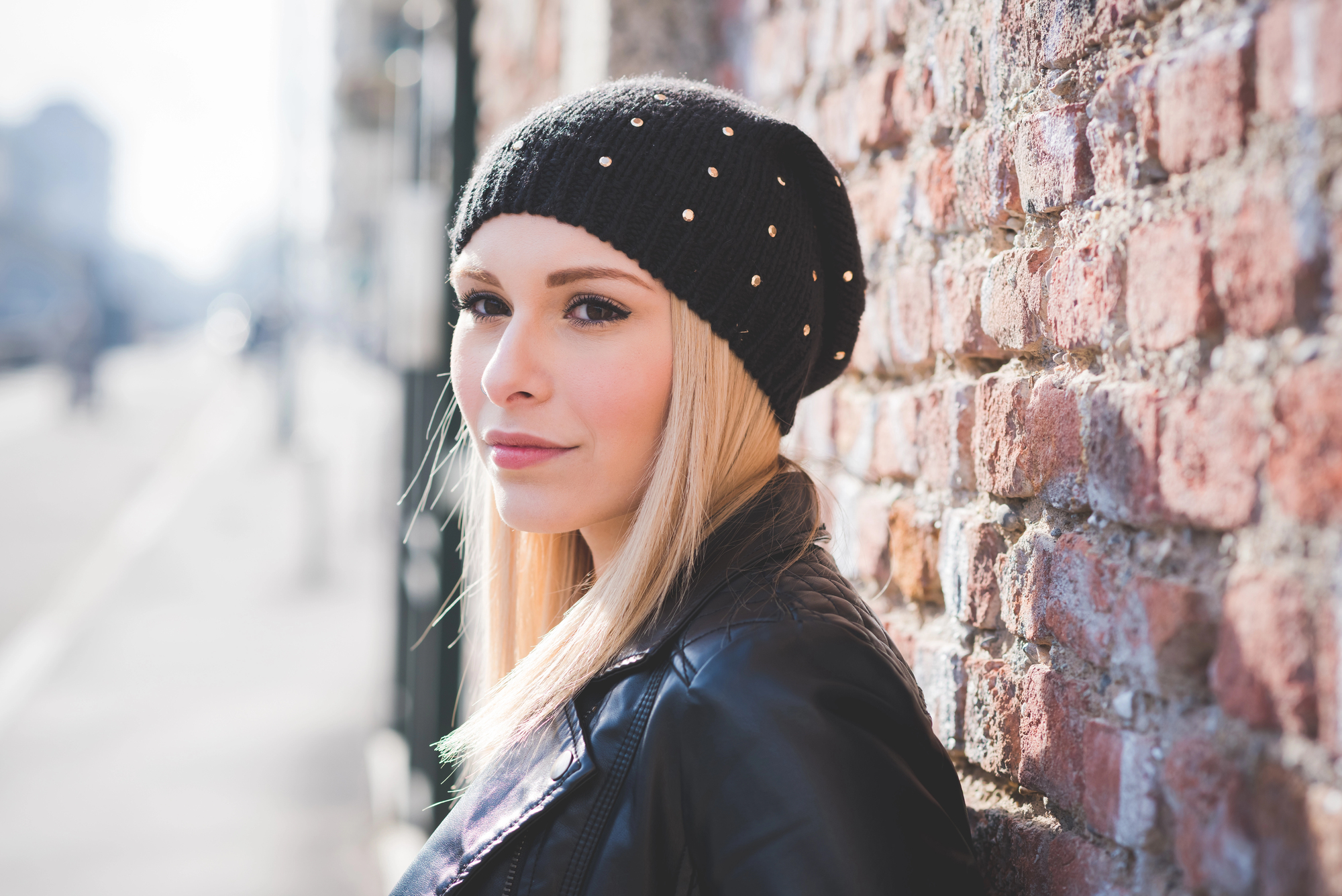 A woman with long blonde hair wearing a black beanie and leather jacket leans against a brick wall. She gazes sideways with a calm expression. The background is blurred, suggesting an urban setting.