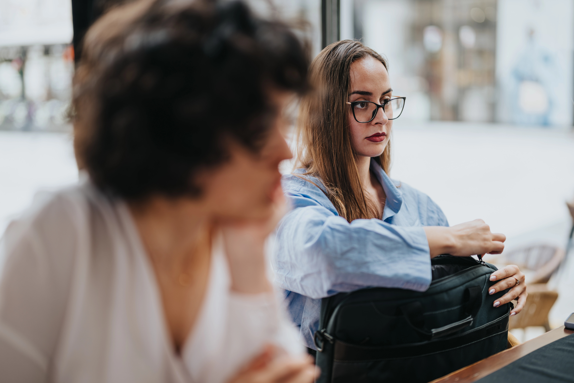 Two women are seated indoors. The woman in focus, wearing glasses and a blue blouse, is looking at the other woman while reaching into her bag. The other woman is out of focus in the foreground, facing away from the camera.