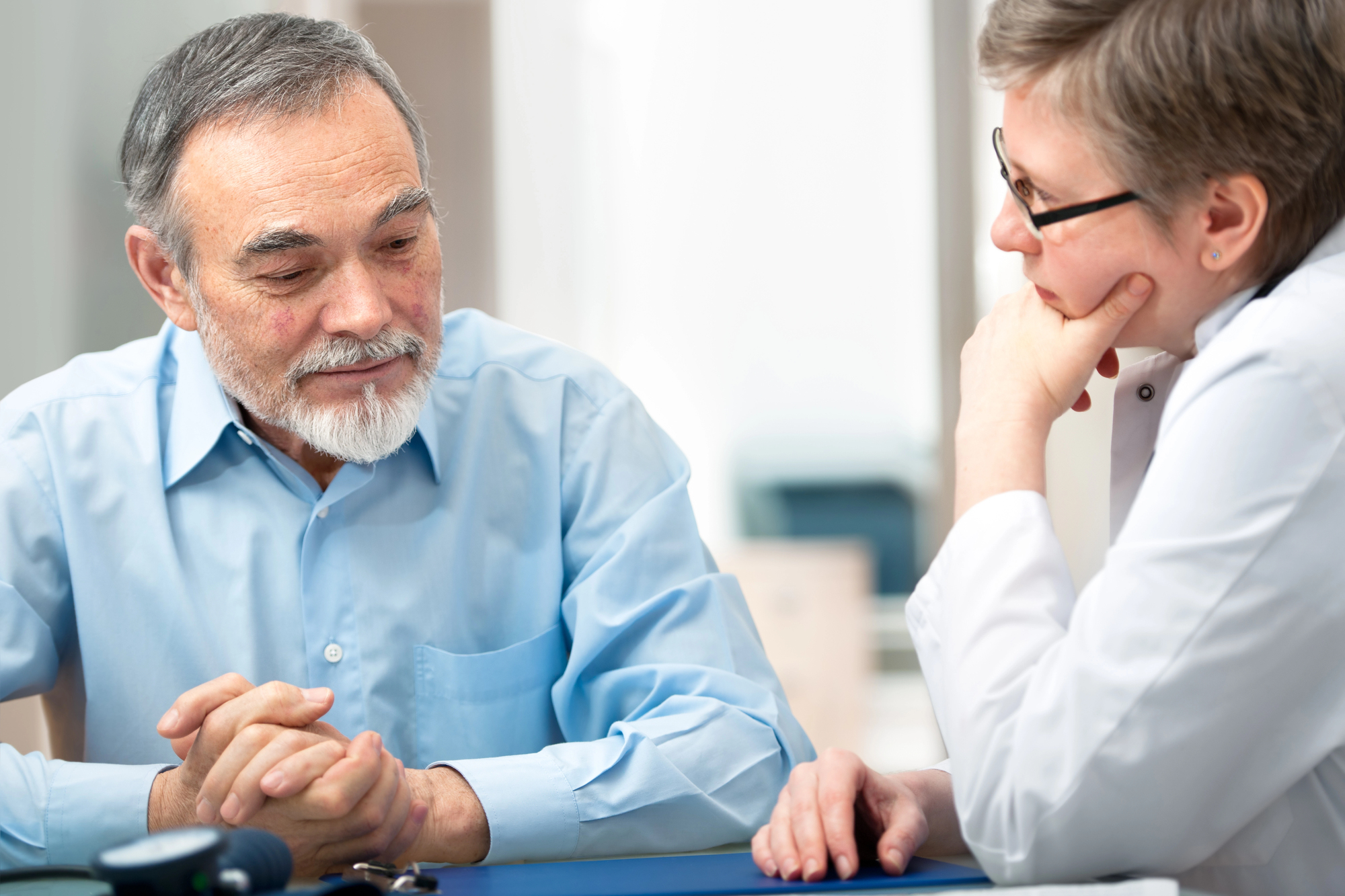 An elderly man with a beard listens attentively during a conversation with a healthcare professional. The healthcare professional is wearing glasses and a white coat, resting her chin on her hand. They are sitting at a desk in a well-lit room.