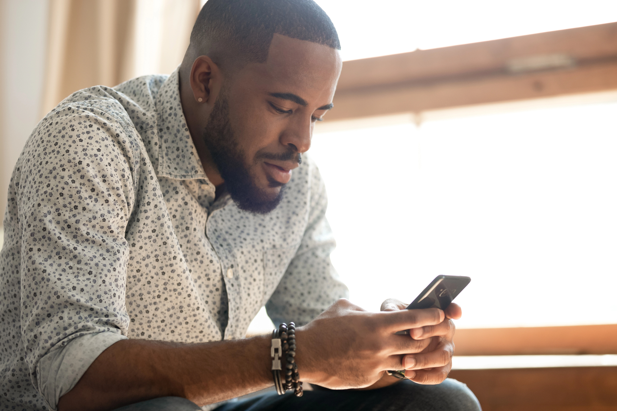 A man with a beard is sitting by a window, wearing a light-colored patterned shirt and bracelets. He is smiling and looking at his smartphone. The sunlight is softly illuminating the background.