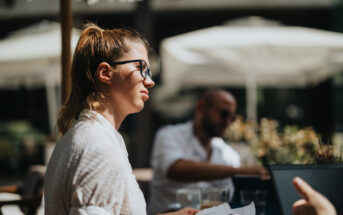 A woman with glasses and a ponytail sits outdoors at a table, looking to the side. She holds papers, surrounded by bright sunlight and blurred background figures. The setting appears to be a casual gathering or meeting.