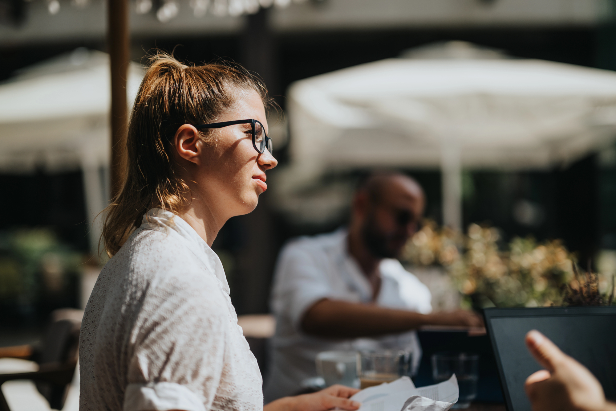 A woman with glasses and a ponytail sits outdoors at a table, looking to the side. She holds papers, surrounded by bright sunlight and blurred background figures. The setting appears to be a casual gathering or meeting.