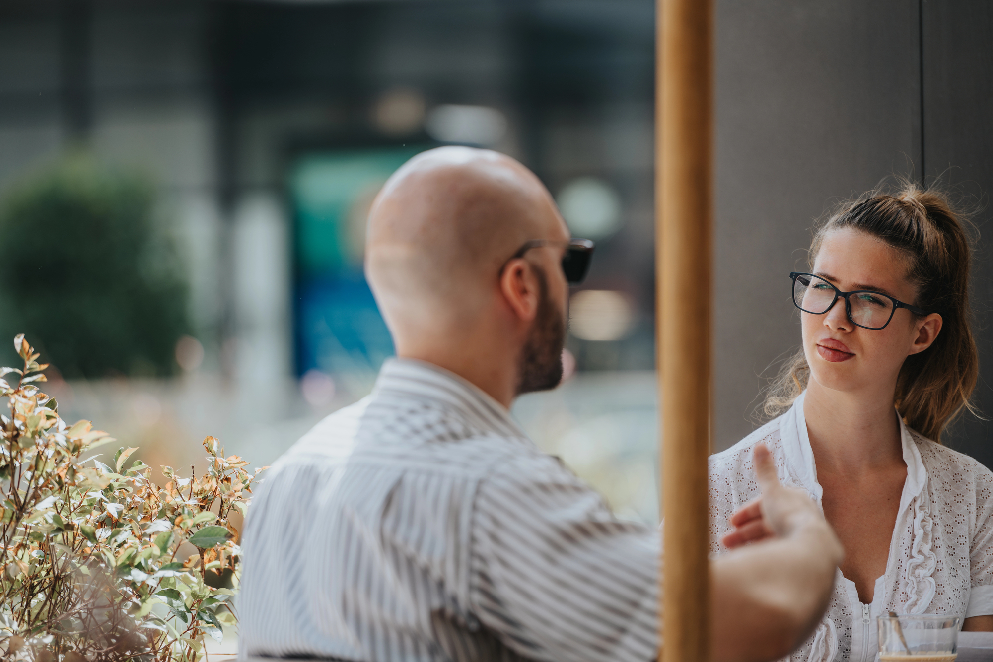 A man and woman sit at an outdoor café table engaged in conversation. The man has a shaved head and wears sunglasses, while the woman has glasses and her hair in a ponytail. Sunlight illuminates the scene, with greenery visible in the background.