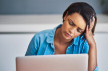 A woman sitting at a desk with her hand on her forehead, looking tired or stressed while staring at a laptop screen. She is wearing a blue button-up shirt.