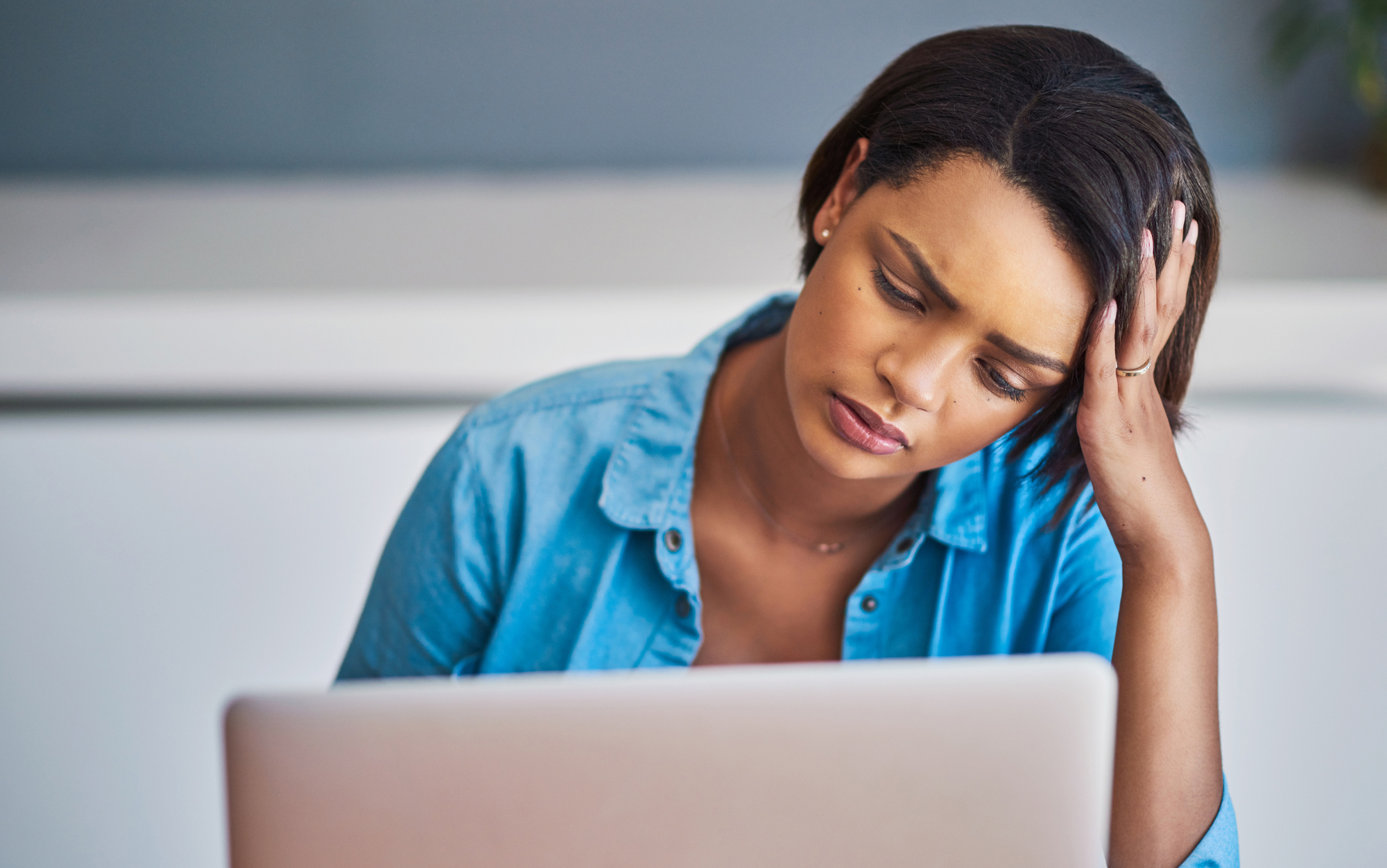 A woman sitting at a desk with her hand on her forehead, looking tired or stressed while staring at a laptop screen. She is wearing a blue button-up shirt.