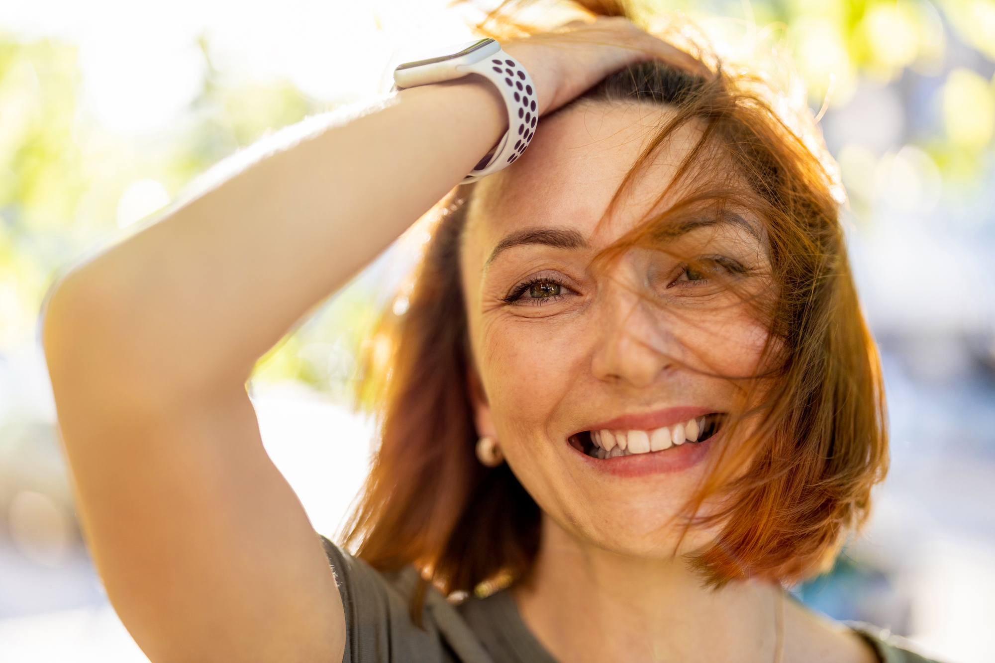 A woman with shoulder-length hair smiles brightly outdoors, her hand running through her hair. Sunlight illuminates her face against a blurred, leafy background, creating a warm and cheerful atmosphere.