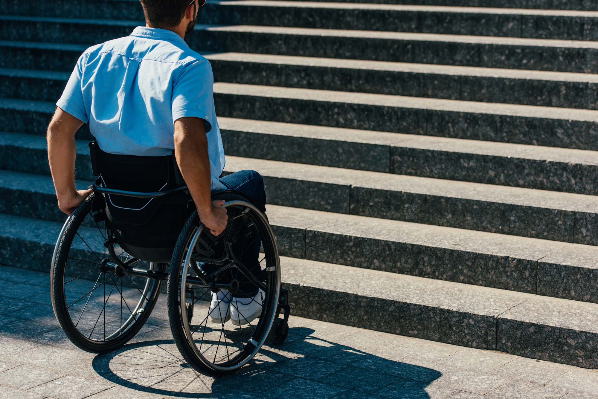 A person in a wheelchair facing a long flight of concrete stairs. The individual is in a blue shirt and jeans, positioned on a stone-paved pathway. The image highlights accessibility challenges.