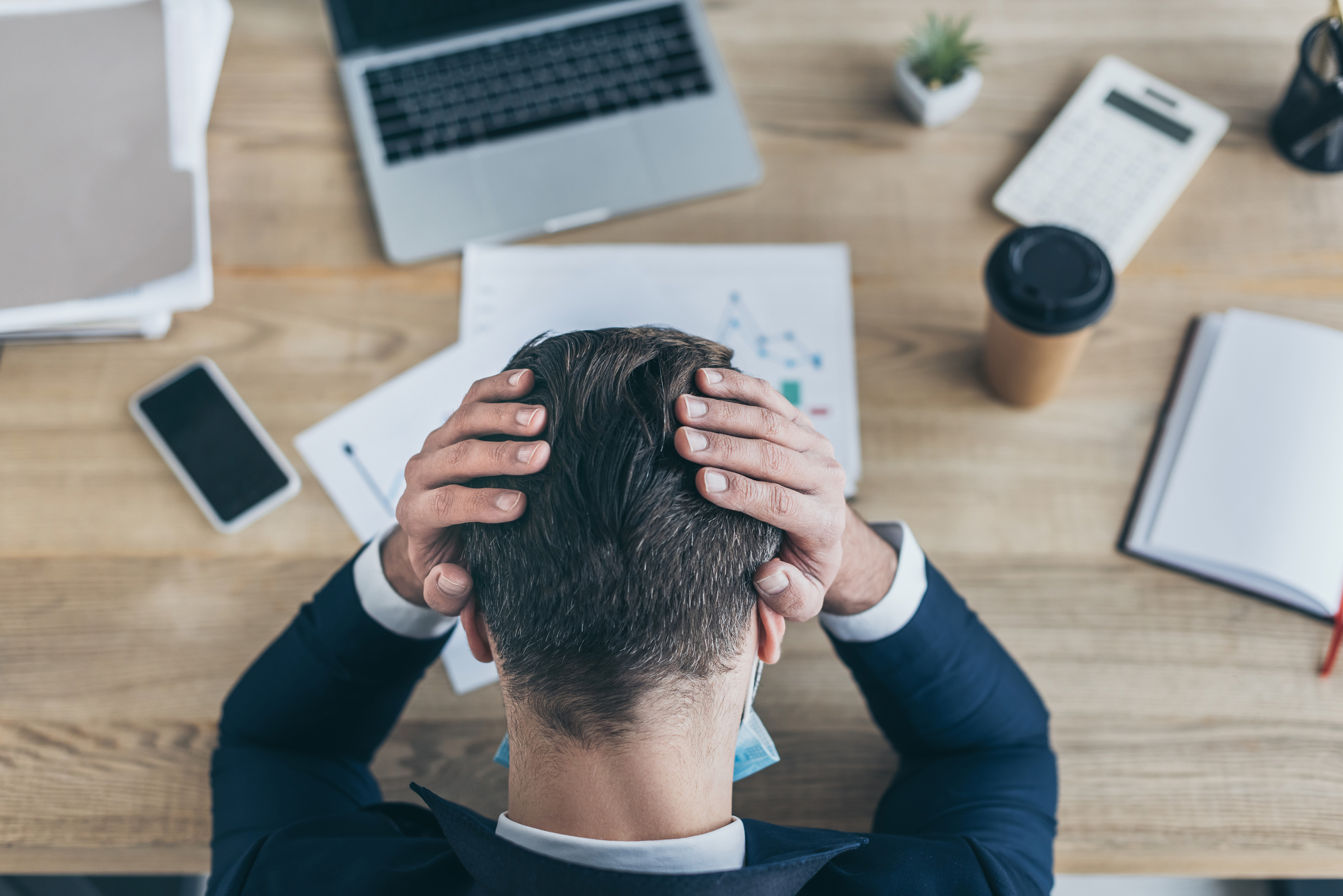 A man in a suit sits at a wooden desk with his hands on his head, looking stressed. The desk has a laptop, documents, a smartphone, a calculator, a notebook, and a coffee cup, with a small plant in the background.