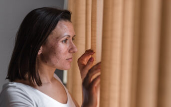 A woman with medium-length brown hair looks out through beige curtains, her expression contemplative and serious. She wears a white shirt and gently touches the fabric with one hand. The lighting is soft and natural.