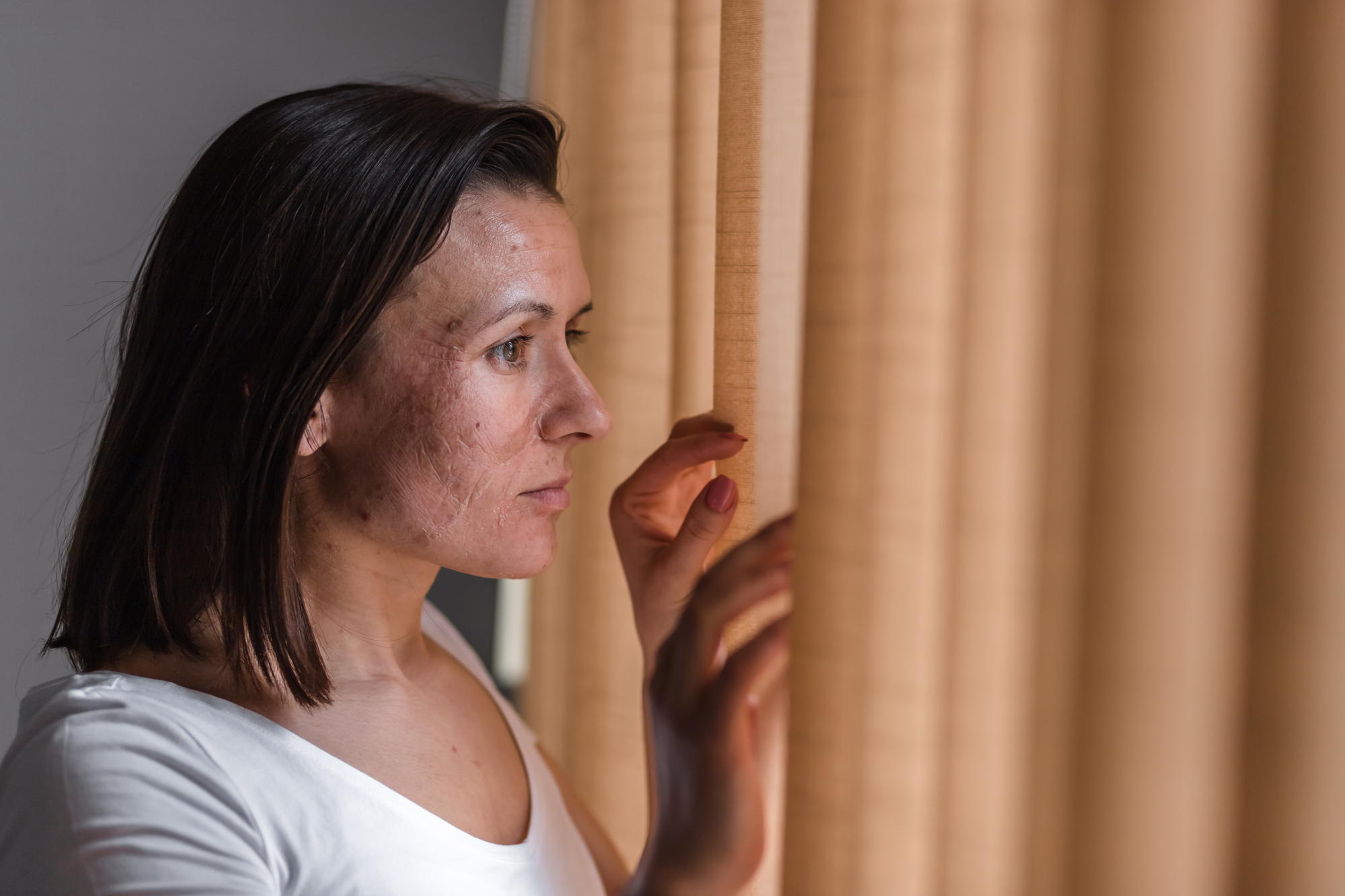 A woman with medium-length brown hair looks out through beige curtains, her expression contemplative and serious. She wears a white shirt and gently touches the fabric with one hand. The lighting is soft and natural.