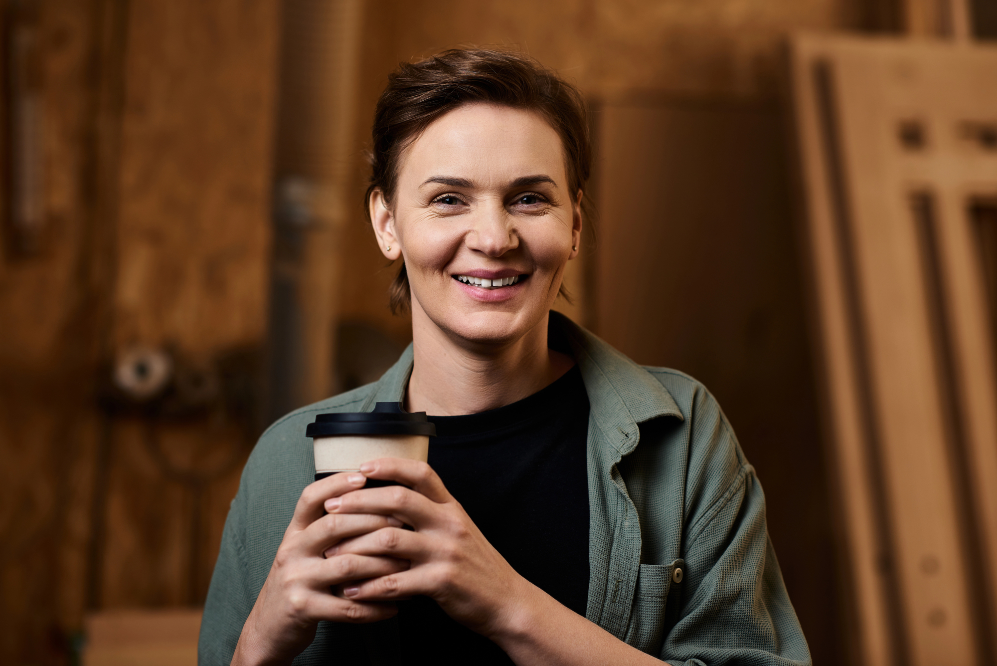 A person smiling and holding a to-go coffee cup with both hands. They are wearing a green jacket over a black shirt. The background is a workshop with tools and wooden materials.