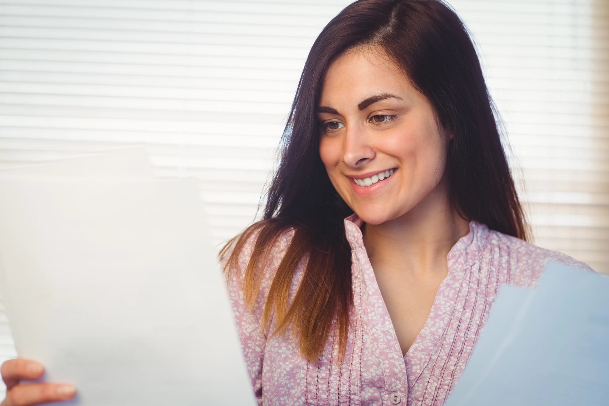 A woman with long dark hair wearing a light pink blouse is smiling while holding and looking at several sheets of paper. She is standing in front of a window with blinds.