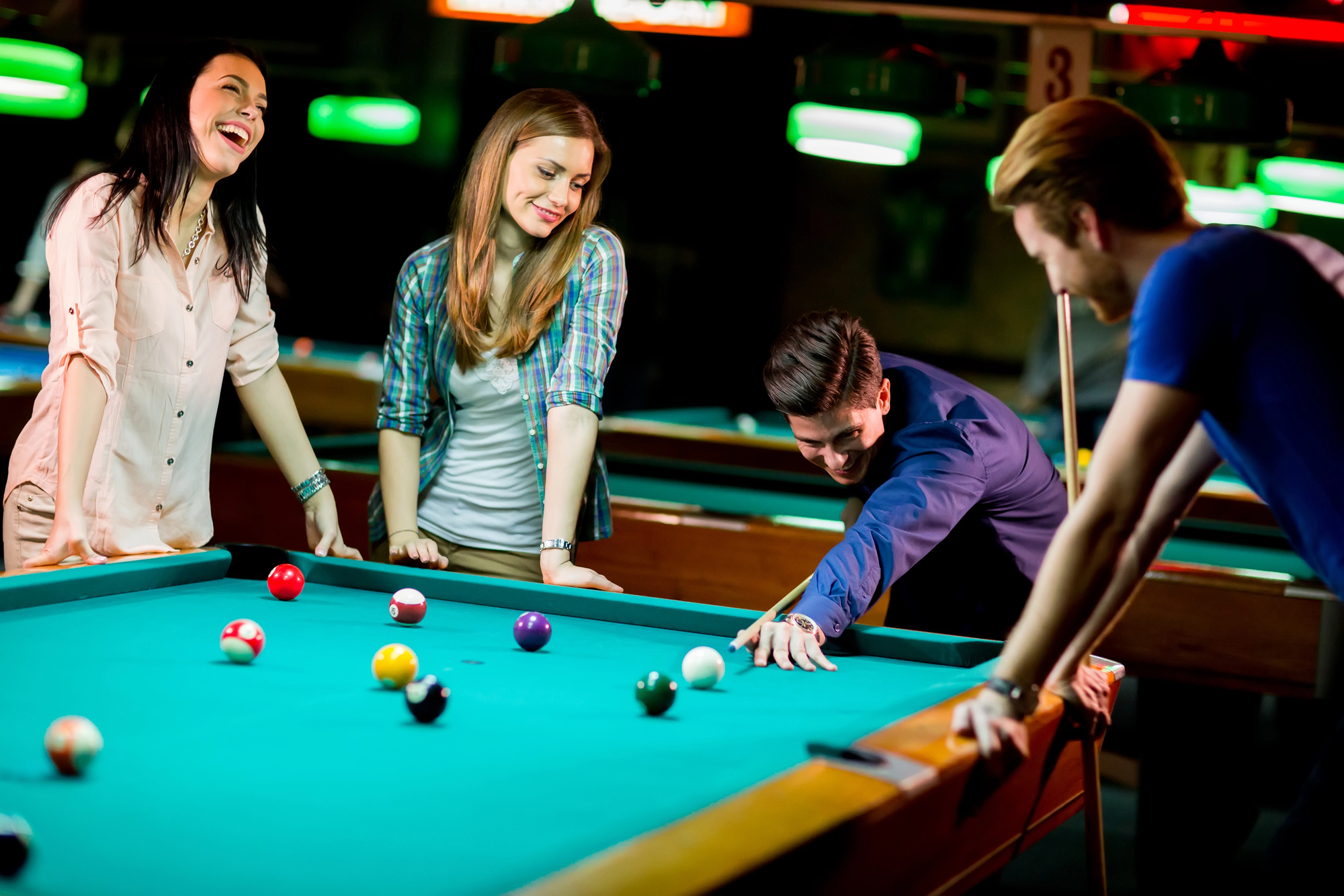 Four friends enjoy a game of pool in a dimly lit bar. One person is taking a shot while the others watch and laugh. The pool table is scattered with balls, and the atmosphere is lively and relaxed.