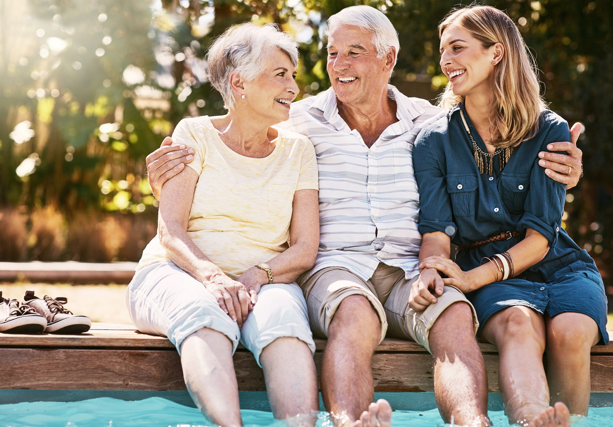 Three people sit together on the edge of a pool, smiling and laughing. An older couple, a man and woman, are sitting next to a younger woman. They are enjoying a sunny day, with their feet in the water. Casual clothing and trees in the background.