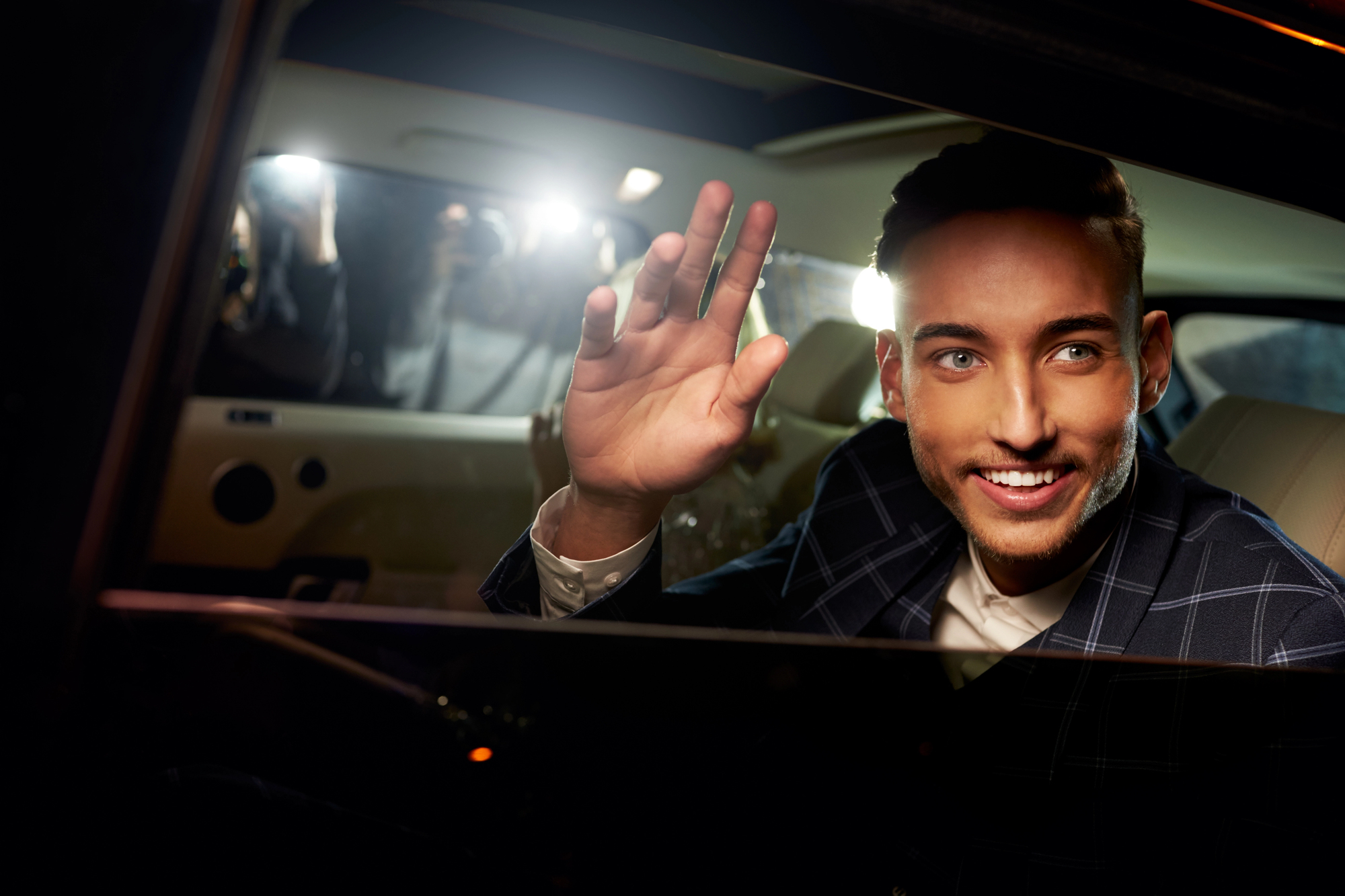 A man in a suit smiles and waves from inside a car as flashes from outside illuminate him. The interior is softly lit, and the window is partially down, allowing him to interact with the onlookers.