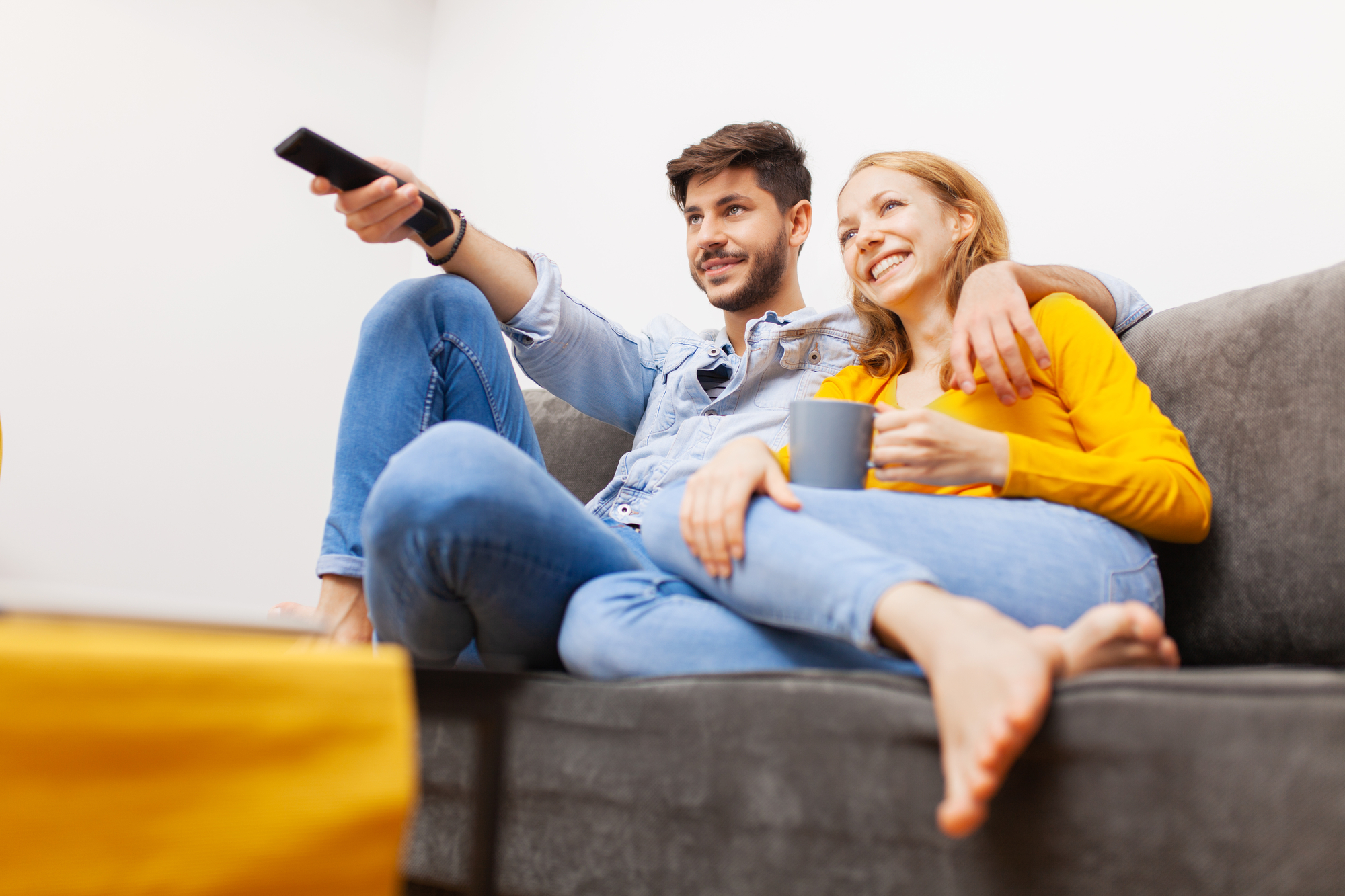 A couple sitting on a gray couch, watching TV. The man, holding a remote, is wearing a denim shirt and jeans. The woman, wearing a yellow sweater and jeans, holds a mug and leans against him, smiling. The setting appears cozy and relaxed.