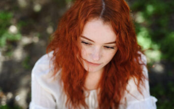 A person with long, curly red hair is looking down thoughtfully. They are wearing a white blouse and are surrounded by a softly blurred green background, suggesting an outdoor setting.