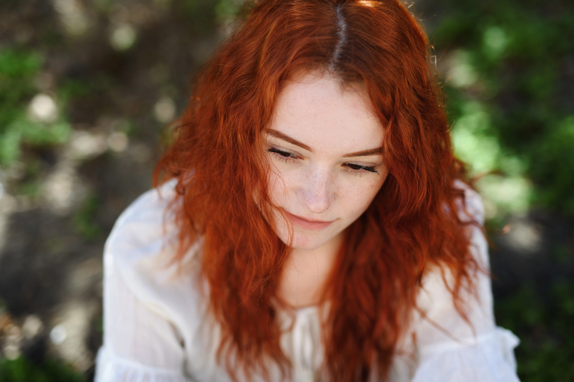 A person with long, curly red hair is looking down thoughtfully. They are wearing a white blouse and are surrounded by a softly blurred green background, suggesting an outdoor setting.