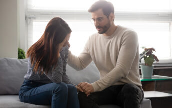 A man in a light sweater consoles a woman sitting beside him on a couch. The woman has her head down and appears distressed. He gently touches her shoulder, offering support. A lamp and potted plant are in the background.