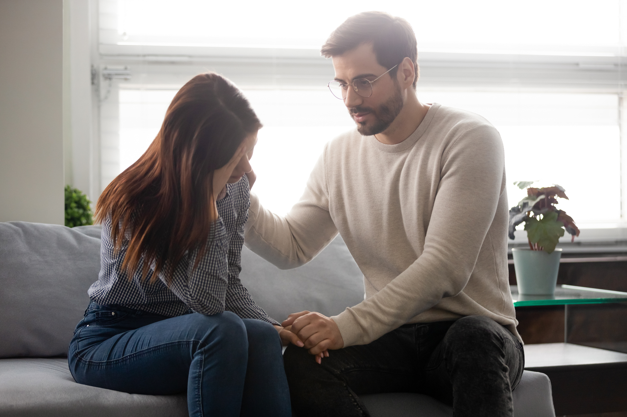 A man in a light sweater consoles a woman sitting beside him on a couch. The woman has her head down and appears distressed. He gently touches her shoulder, offering support. A lamp and potted plant are in the background.