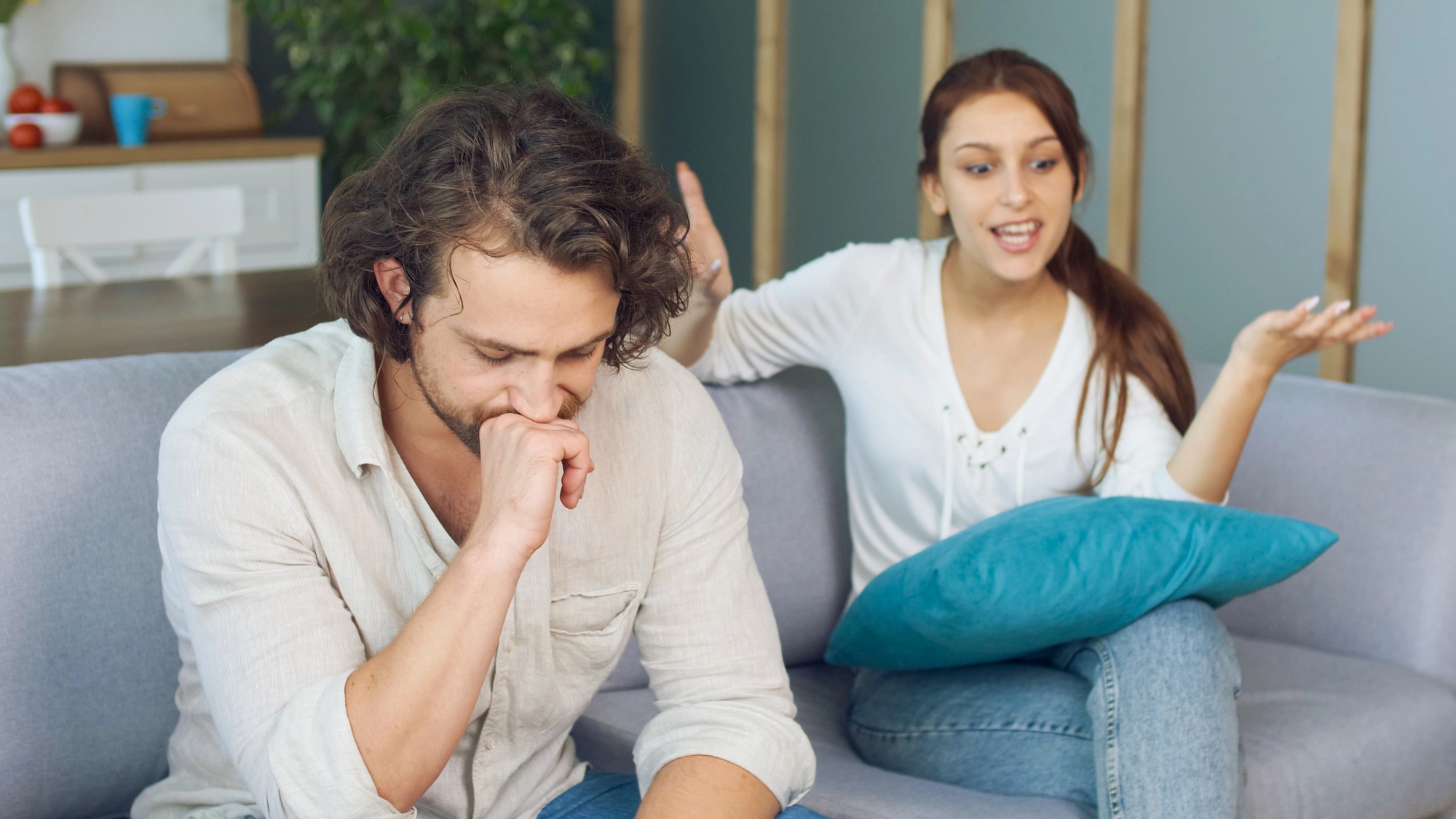 A man and woman are sitting on a couch. The man looks thoughtful, resting his chin on his hand. The woman is talking energetically, gesturing with her hands. A blue pillow is on her lap. The setting is a modern living room.