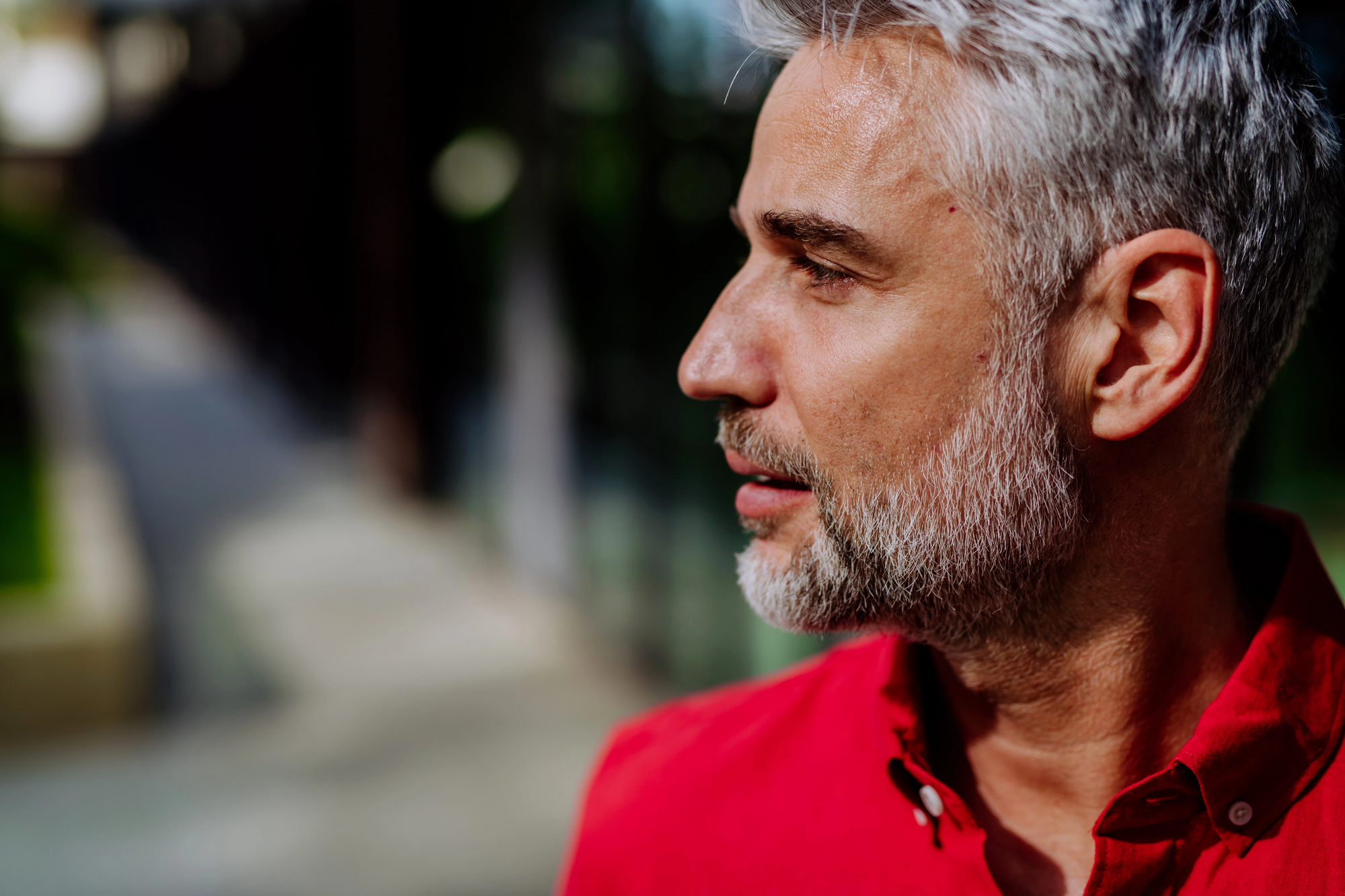 A man with gray hair and a beard wearing a red shirt stands outdoors, looking to the side. The background is blurred, featuring a walkway and greenery.