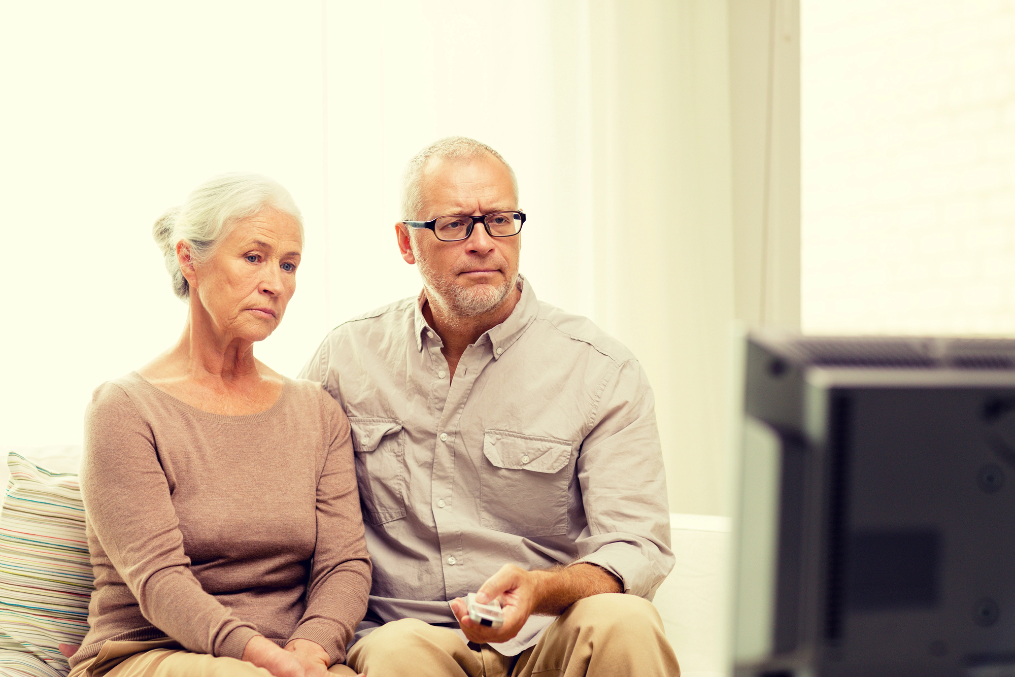 An elderly couple sits on a couch watching TV. The man holds a remote control, and both appear focused on the screen. The room is softly lit, with a white wall and a window in the background. They wear casual, comfortable clothing.