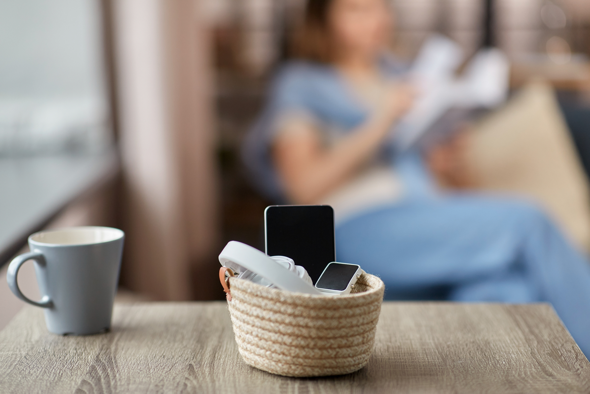 A woven basket with a smartphone, smartwatch, and headphones sits on a wooden table. A gray mug is nearby. In the blurred background, a person is sitting and reading a book.