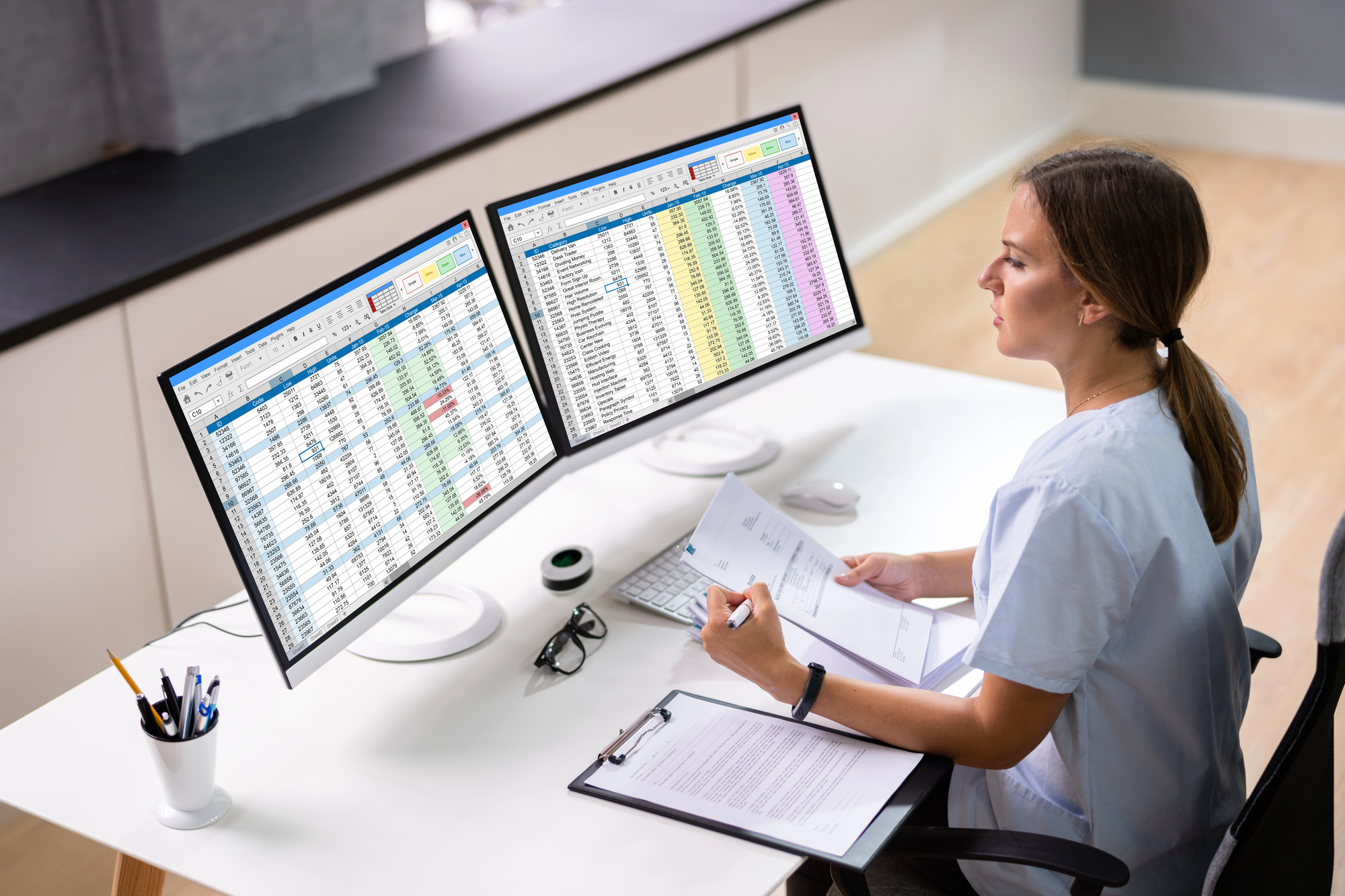 A woman sits at a desk analyzing data on two computer monitors displaying spreadsheets. She holds papers and a pen, with a clipboard and glasses beside her. The workspace is organized, with a pen holder and a minimalistic design.