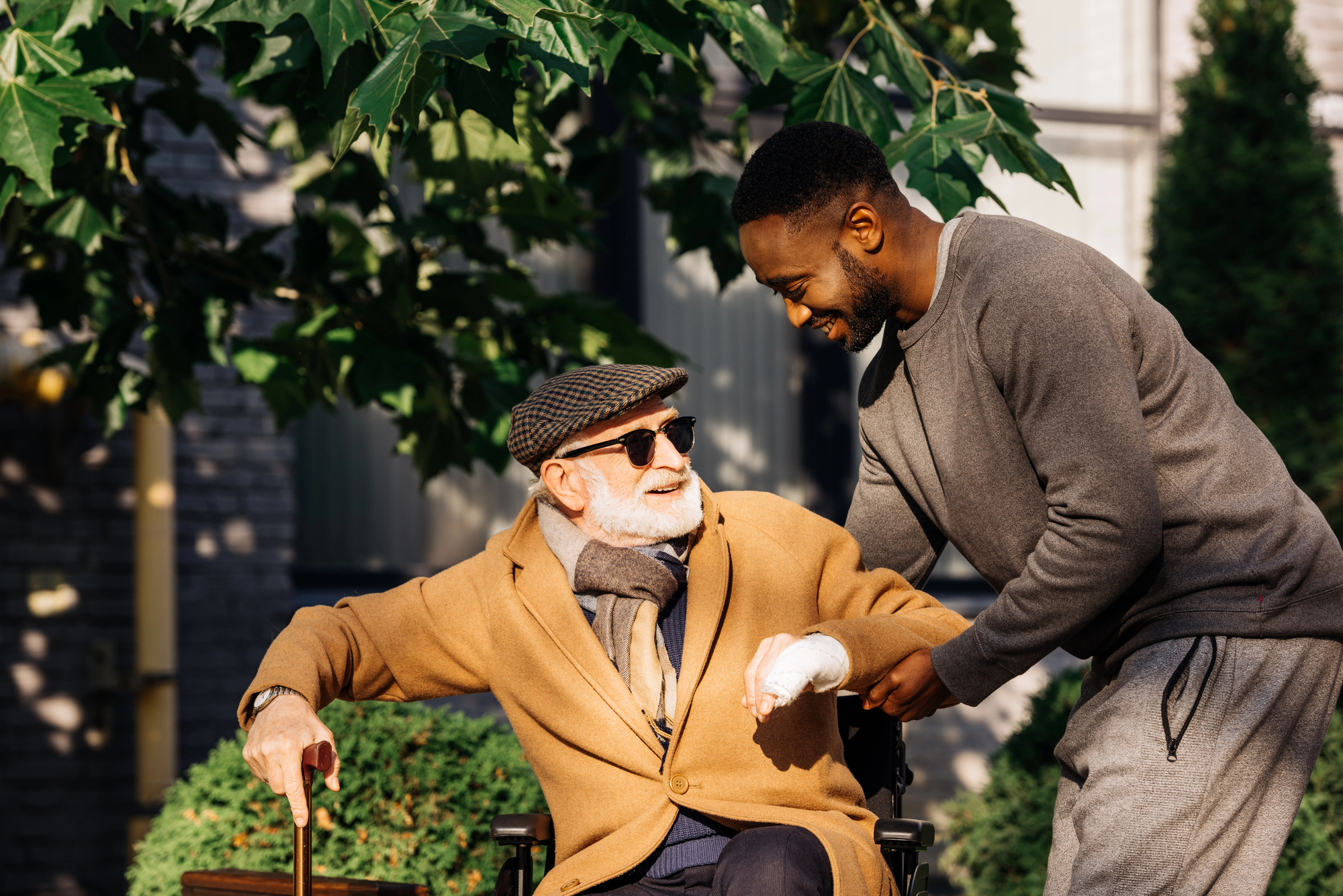 A smiling elderly man in a wheelchair, wearing glasses and a cap, is being assisted by a young man in a sweater. They are outdoors, surrounded by greenery, on a sunny day.