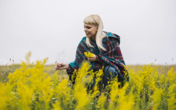 A person with blonde hair and a plaid shawl crouches in a field of tall yellow flowers, smiling as they look at the plants. The sky is overcast, creating a serene, natural setting.