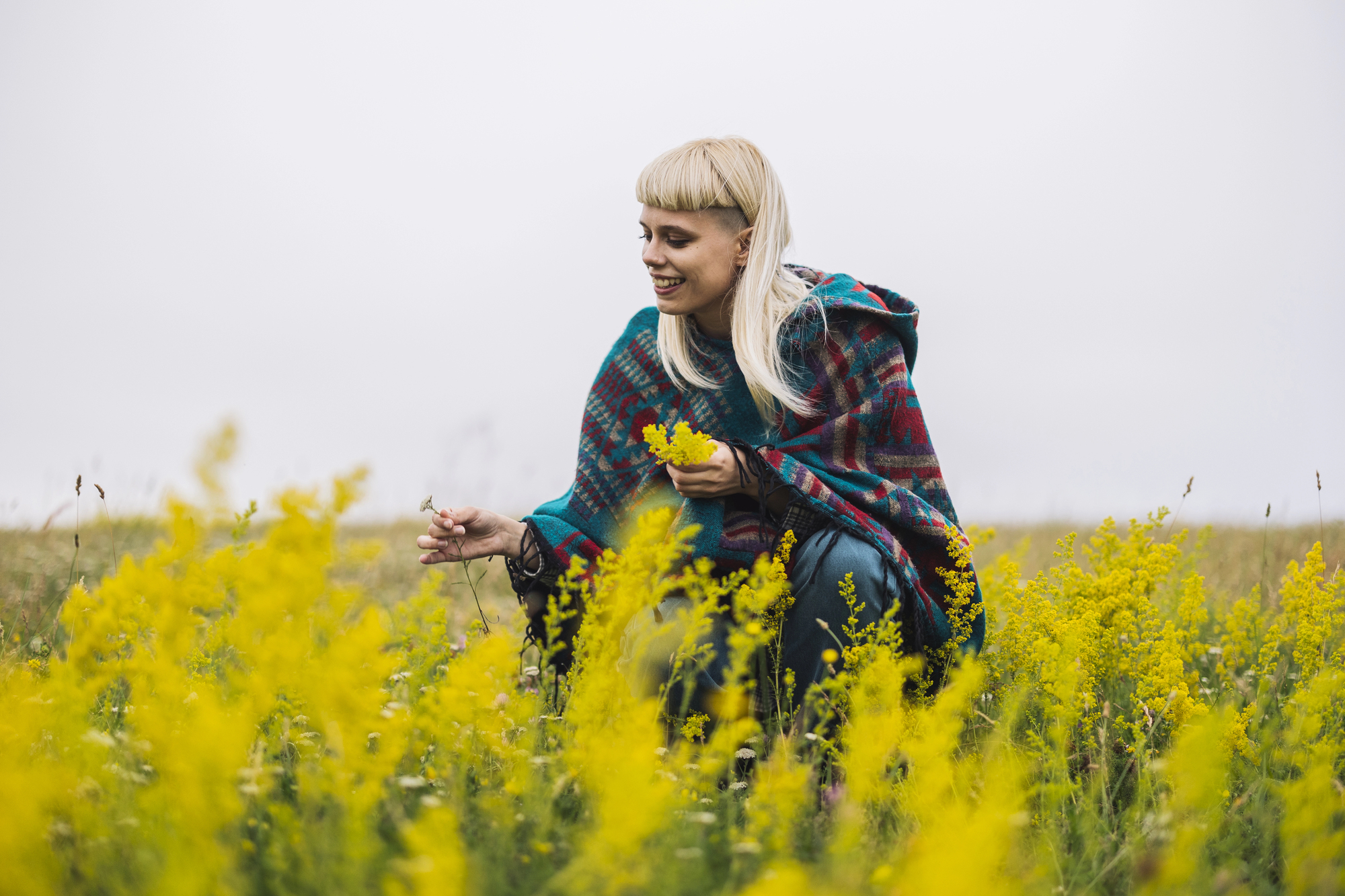 A person with blonde hair and a plaid shawl crouches in a field of tall yellow flowers, smiling as they look at the plants. The sky is overcast, creating a serene, natural setting.