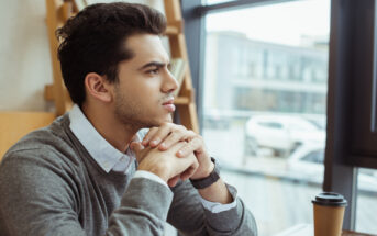 A man with dark hair sits at a table in a cafe, looking thoughtfully out of a window. He rests his chin on his clasped hands. A coffee cup is on the table in front of him, and cars are visible through the window.