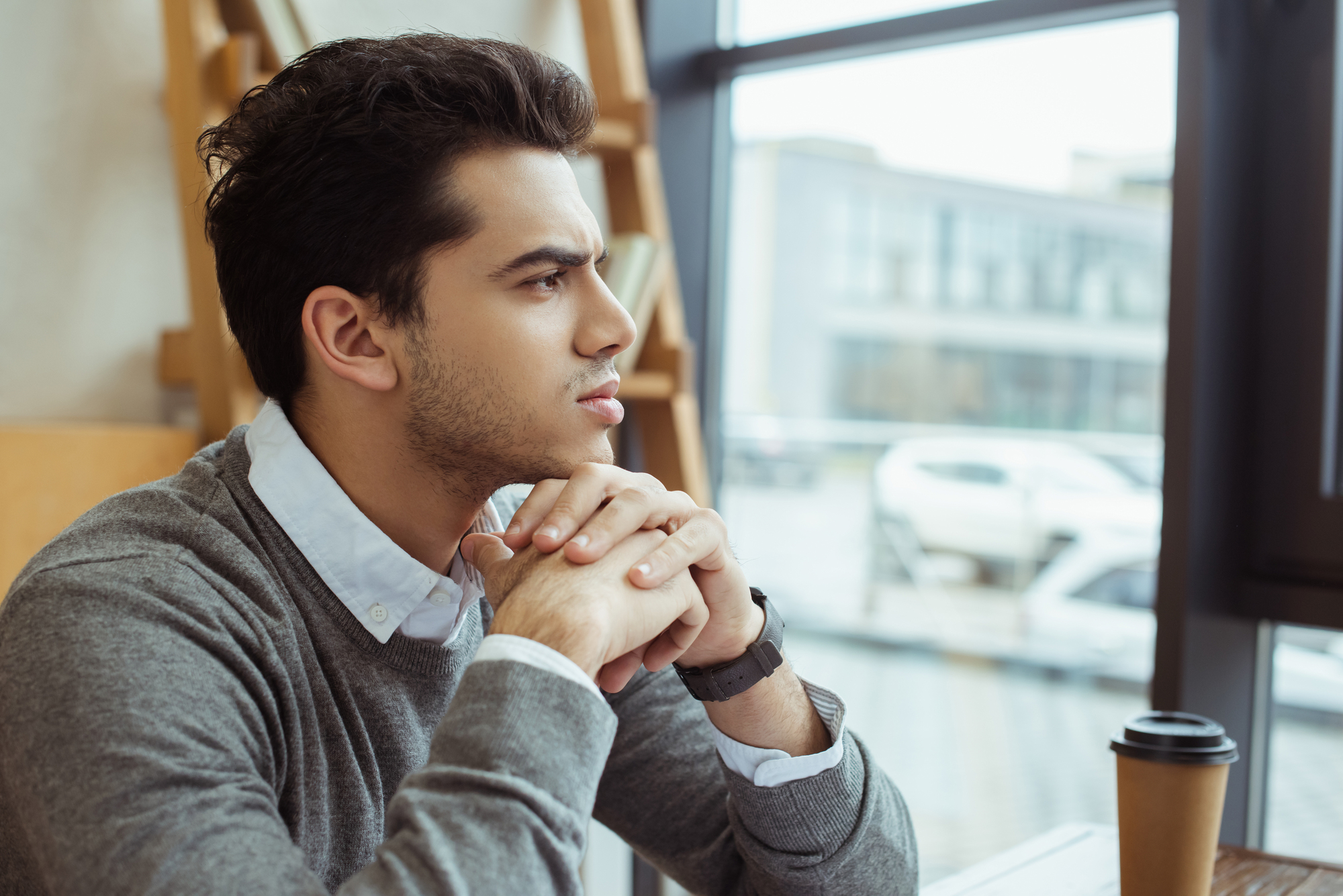 A man with dark hair sits at a table in a cafe, looking thoughtfully out of a window. He rests his chin on his clasped hands. A coffee cup is on the table in front of him, and cars are visible through the window.