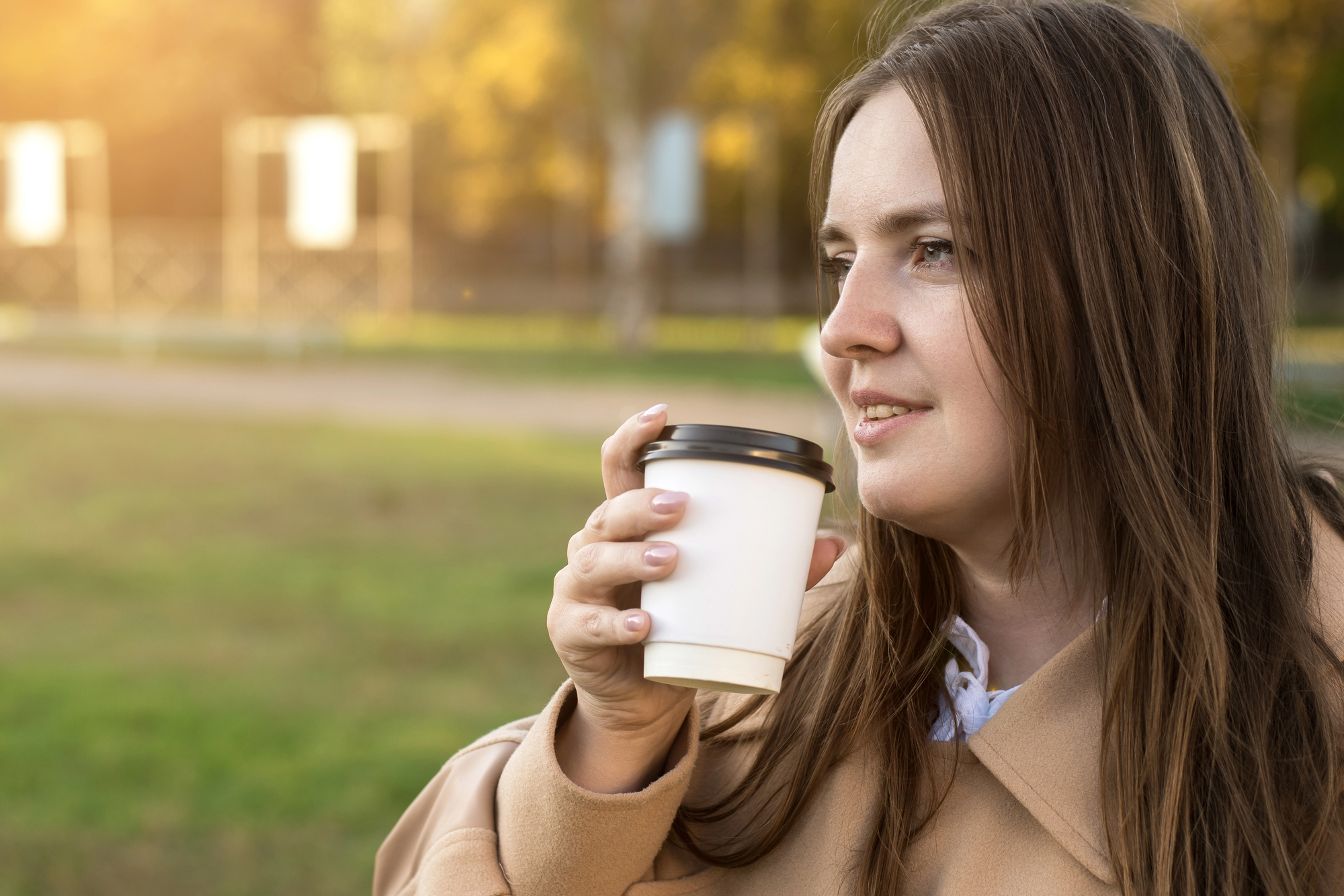 A woman in a beige coat holds a white takeaway coffee cup, smiling slightly and looking into the distance. She's outdoors in a park setting with green grass and blurred trees in the background, bathed in warm sunlight.