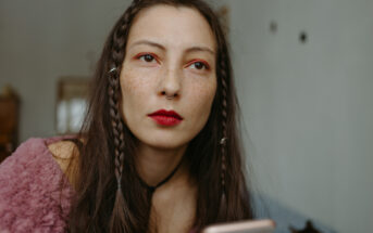 A person with long, braided hair and red lipstick looks intently while holding a smartphone. They are wearing a fuzzy pink top and have a septum piercing. The background is softly blurred.