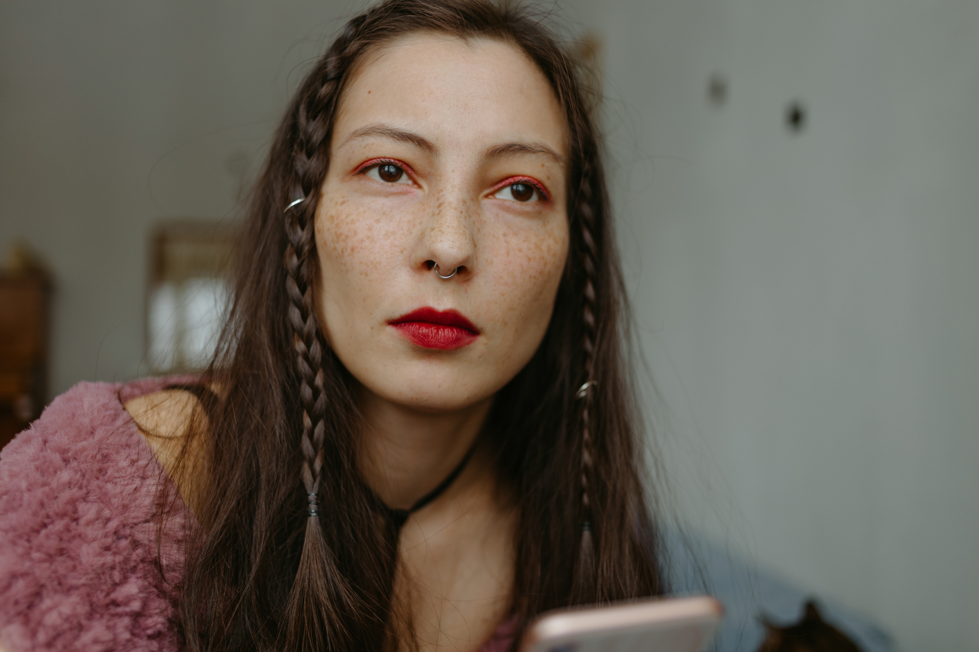 A person with long, braided hair and red lipstick looks intently while holding a smartphone. They are wearing a fuzzy pink top and have a septum piercing. The background is softly blurred.