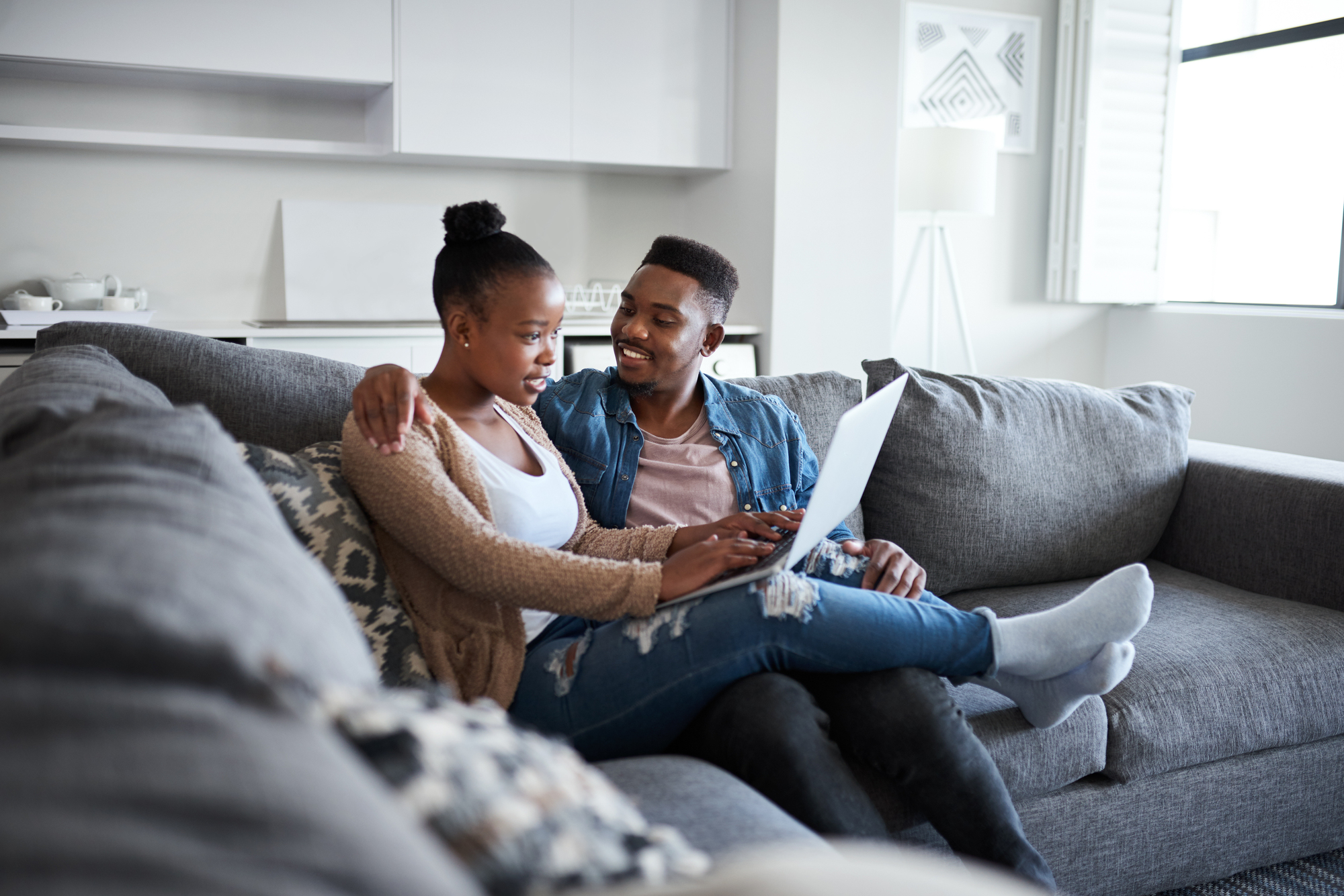 A couple sits comfortably on a gray sofa in a modern living room. The woman is using a laptop on her lap, while the man, sitting beside her, looks on with a smile. The room is bright and decorated with soft colors and patterns.