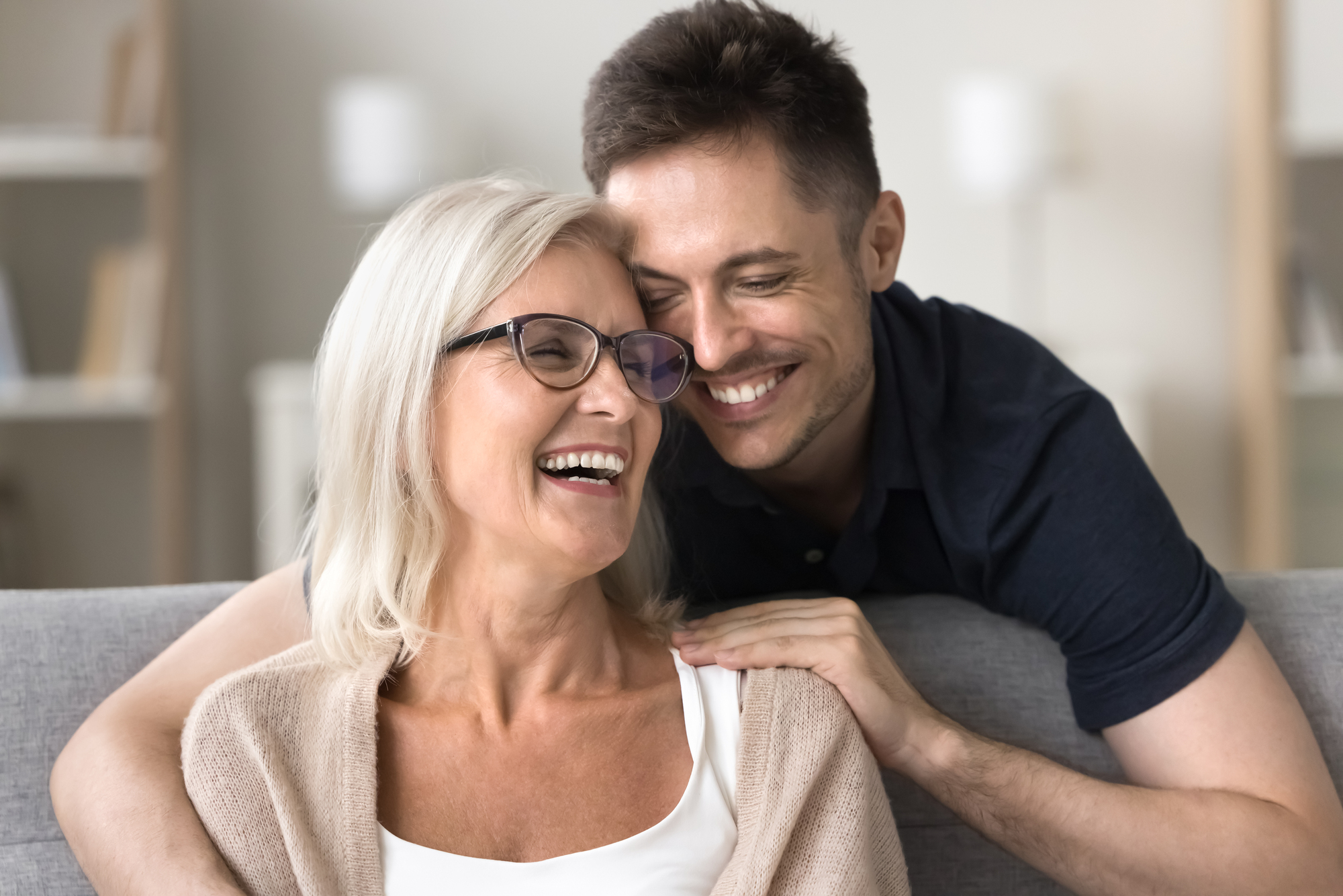 A smiling older woman with glasses and white hair sits on a couch, while a younger man with short brown hair leans affectionately over her shoulder. They both appear happy and relaxed in a cozy living room setting.