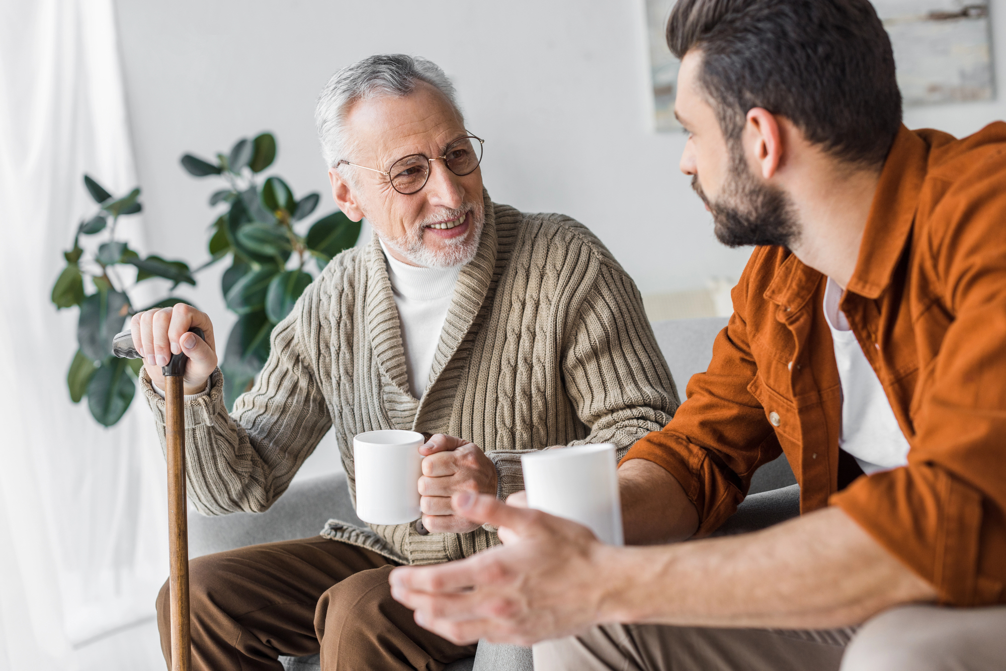 An elderly man with glasses and a cane, wearing a beige cardigan, sits on a couch holding a mug. He is engaged in conversation with a younger man in a brown jacket, who also holds a mug. A leafy plant is in the background.