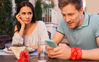 A woman looks at a man with a concerned expression as he smiles while using his smartphone. They are seated at an outdoor table with food and drinks. There is a red flower on the table in the foreground.