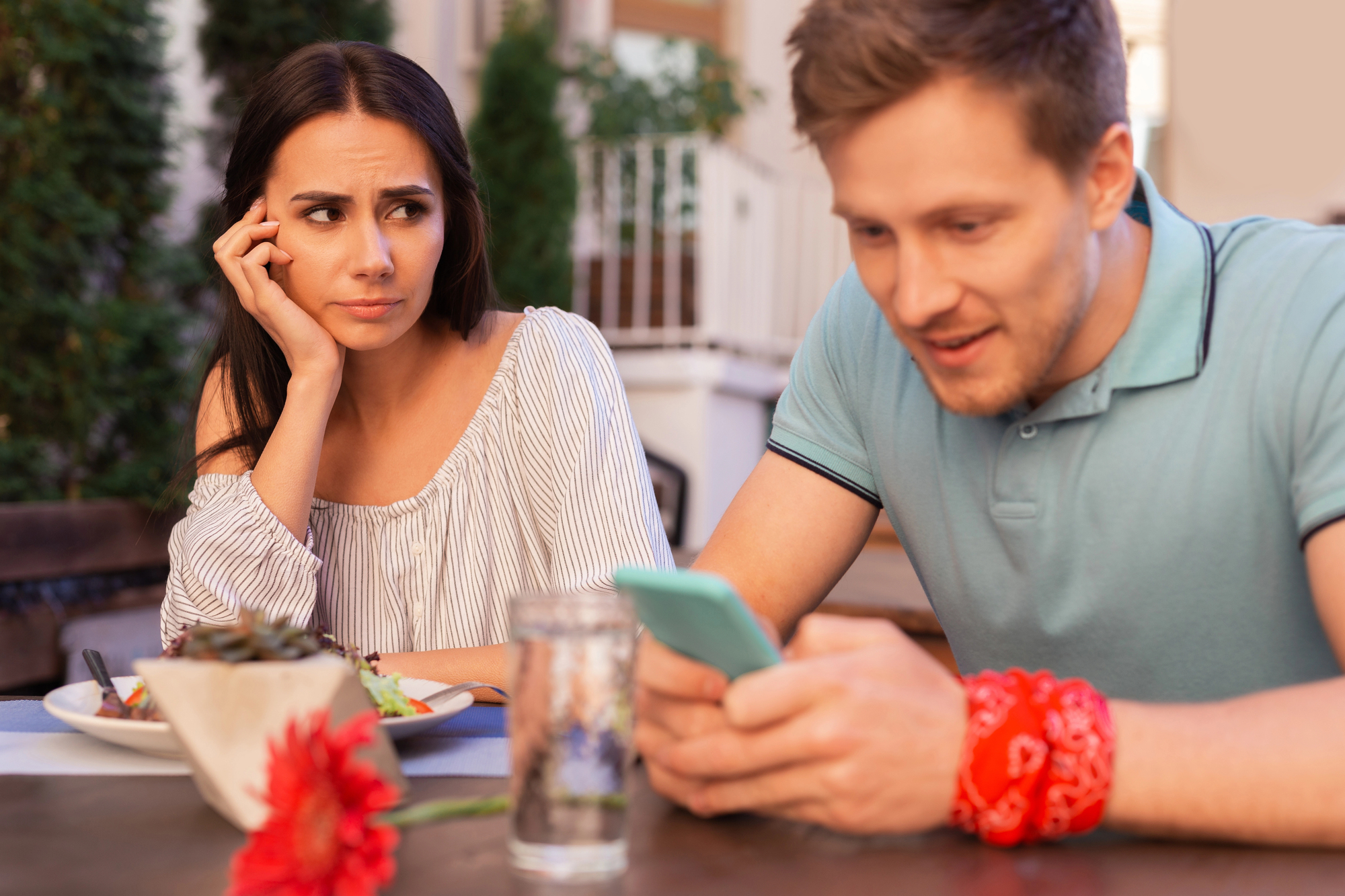 A woman looks at a man with a concerned expression as he smiles while using his smartphone. They are seated at an outdoor table with food and drinks. There is a red flower on the table in the foreground.