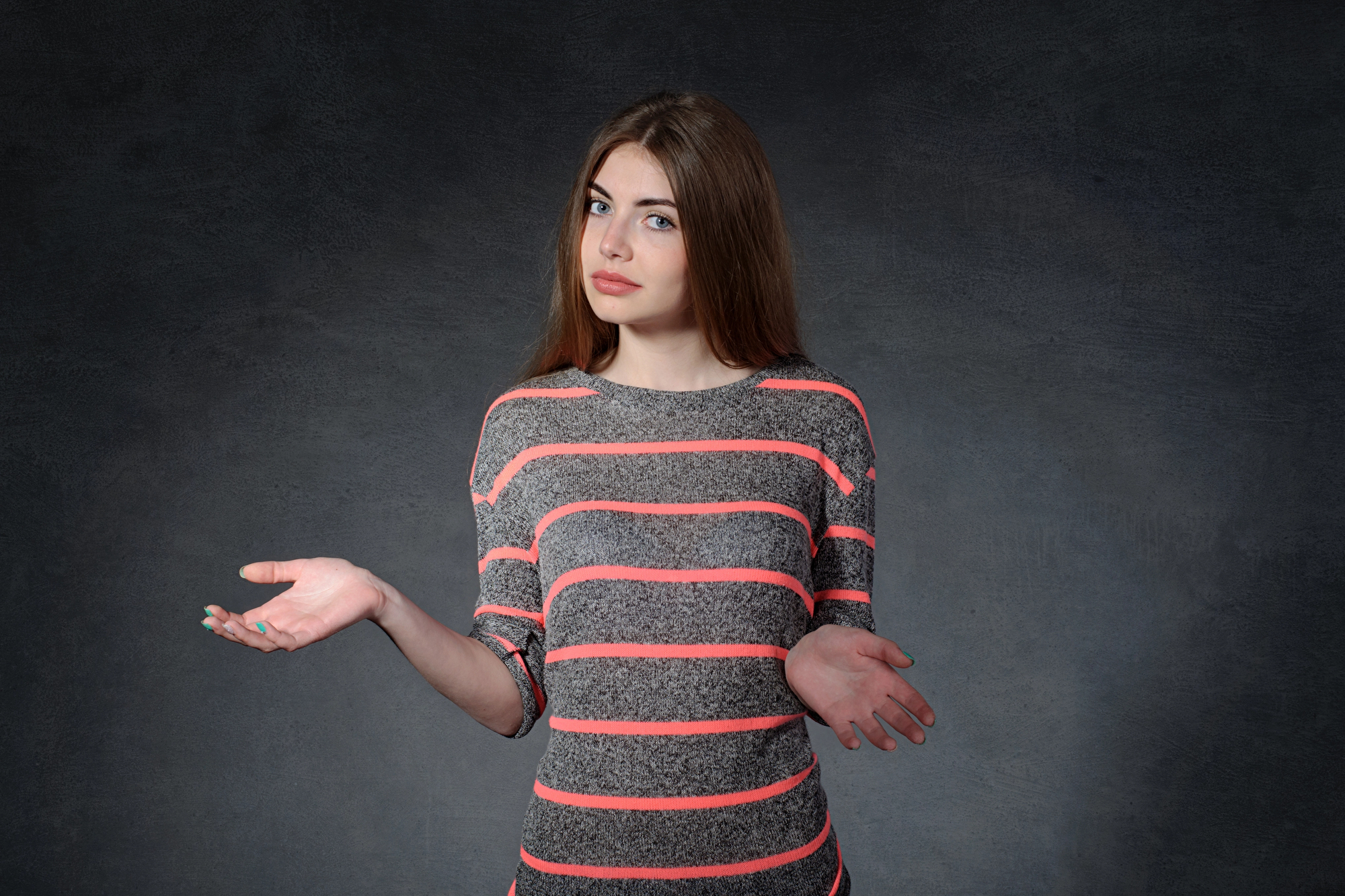 A woman with long brown hair wearing a gray and pink striped sweater gestures with her palms open, standing against a dark gray background. Her expression is neutral, and she appears to be shrugging.