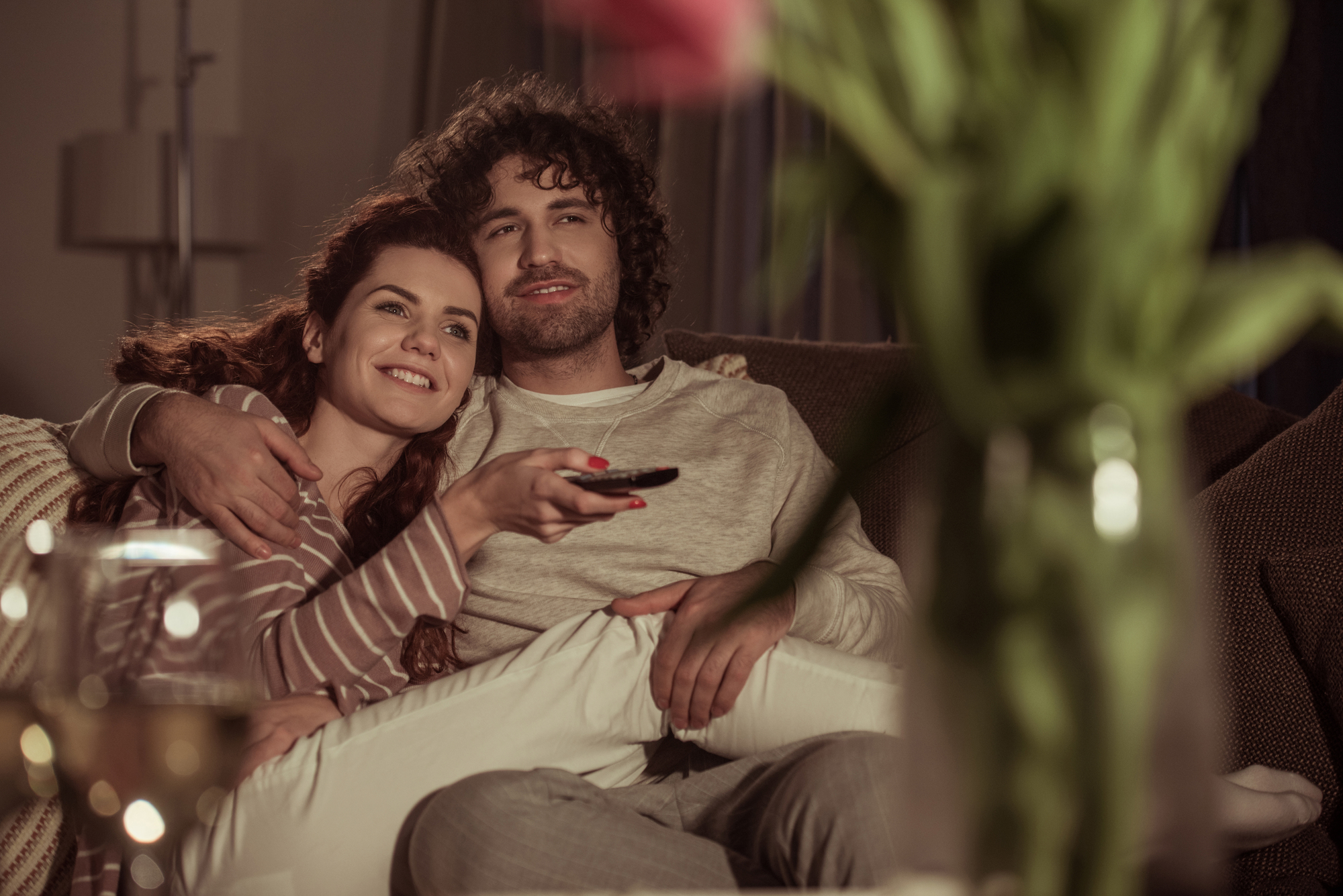 A couple sits closely on a couch, smiling and holding a TV remote. They appear relaxed and cozy, surrounded by soft lighting. A blurred vase with flowers is in the foreground, enhancing the warm, intimate atmosphere.