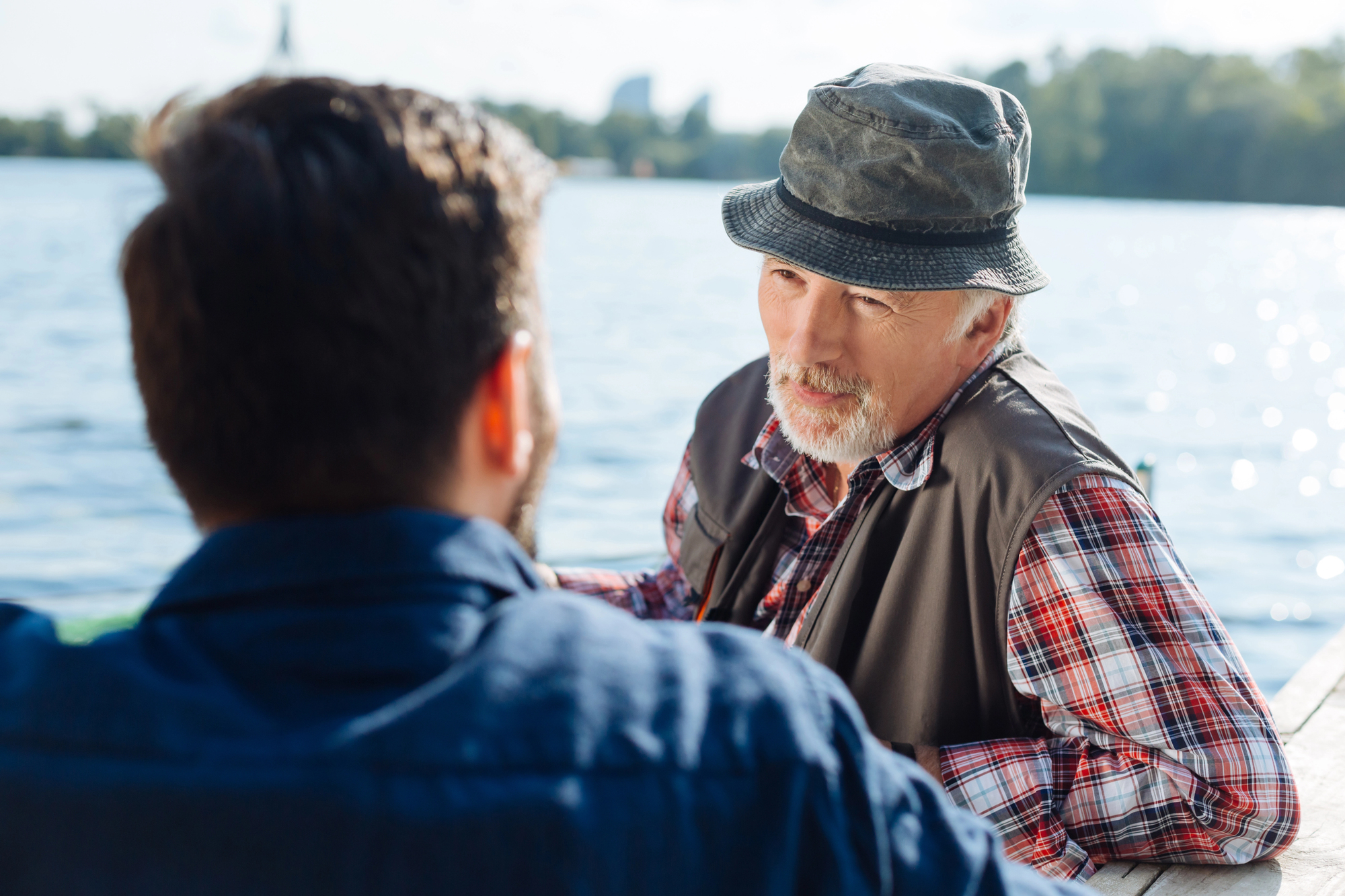 Two men sitting on a pier by a lake, engaged in conversation. The older man, wearing a plaid shirt, vest, and bucket hat, faces forward. The younger man, with dark hair, is seen from behind. Trees and distant buildings are visible across the water.