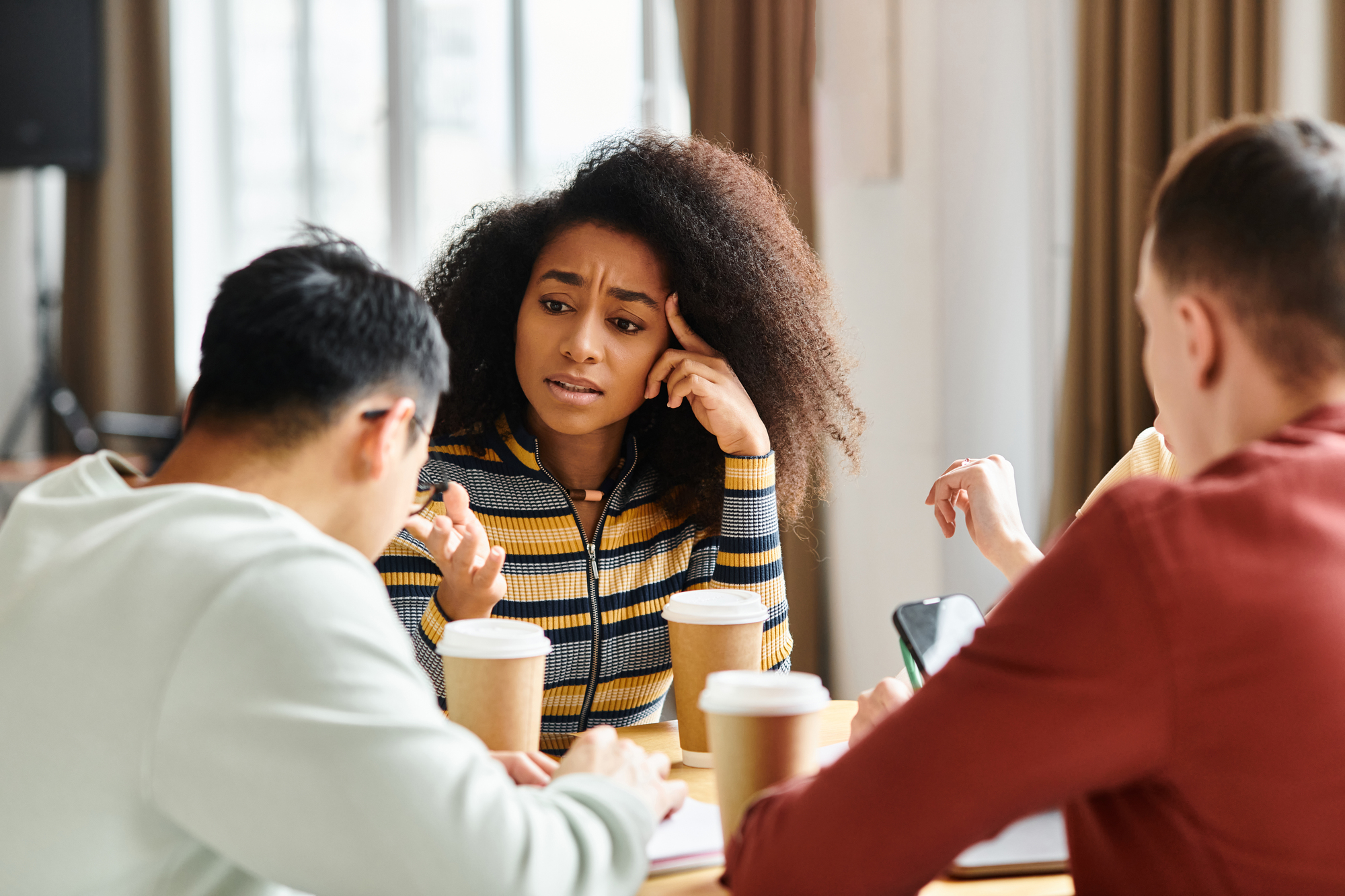 A woman with curly hair gestures expressively while talking to two people seated at a table with coffee cups and a smartphone. The setting appears to be a casual meeting or discussion.