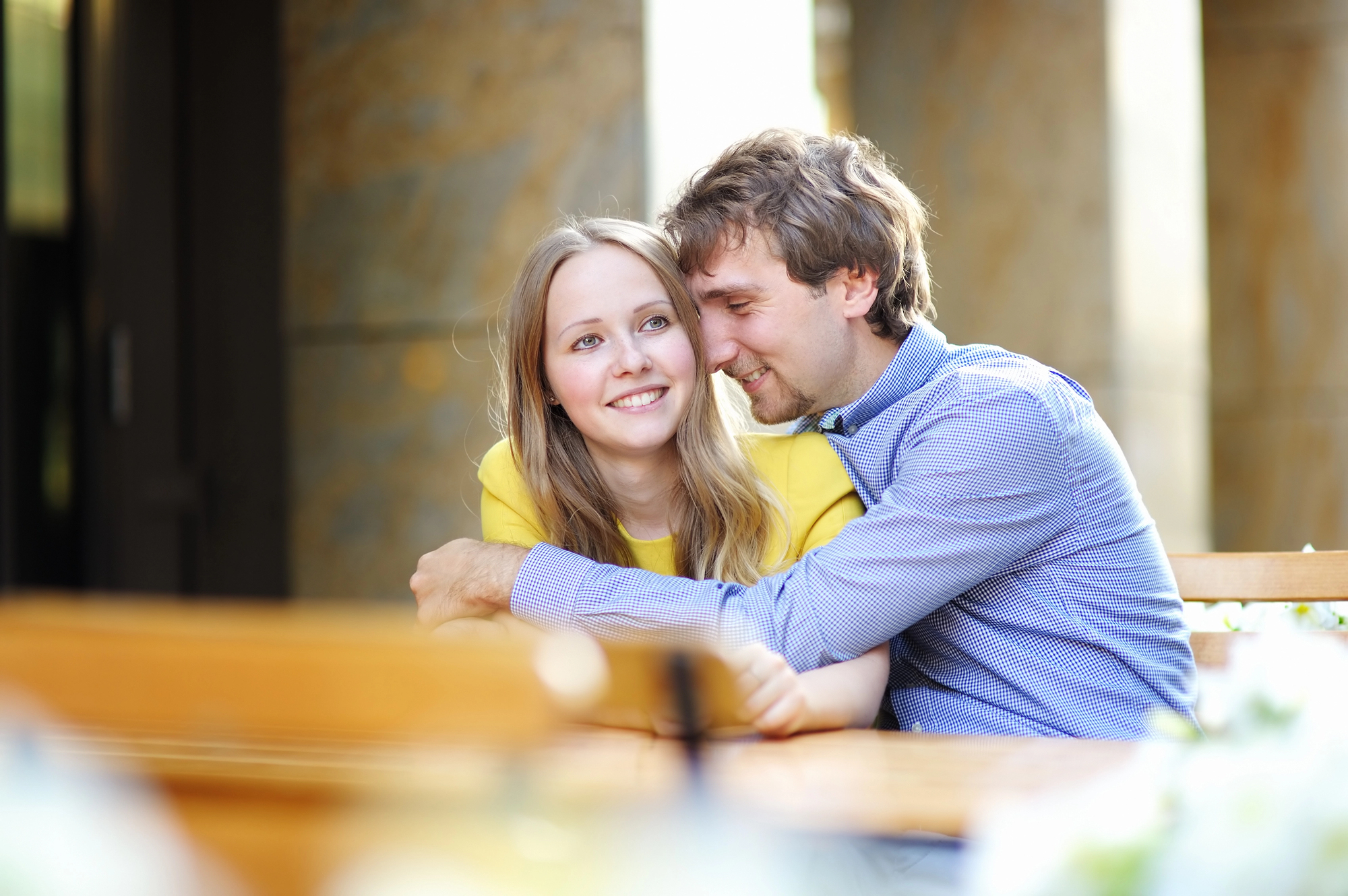 A couple sitting at an outdoor table, smiling and embracing. The woman is wearing a yellow top, and the man is wearing a blue shirt. The background features a soft, blurred view of a building's columns.